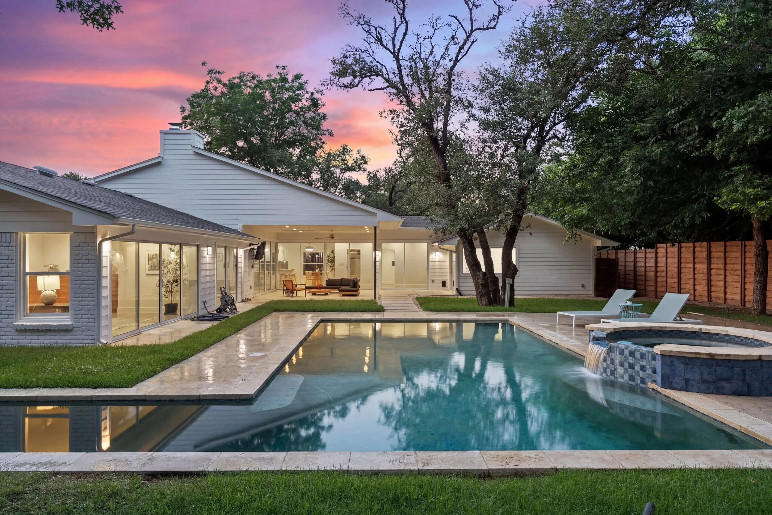 A modern house with a backyard featuring a swimming pool, outdoor seating, and large glass windows at sunset.