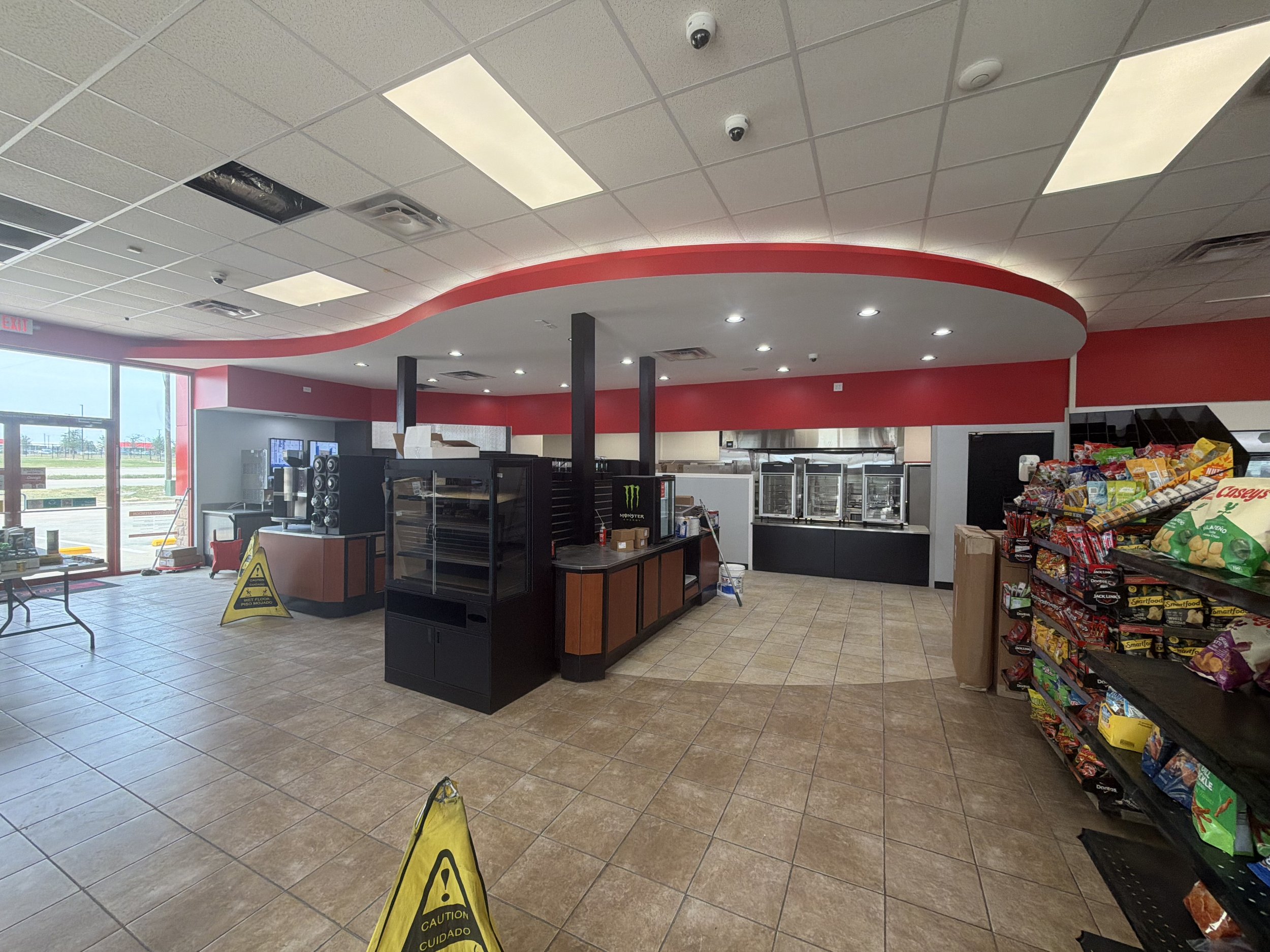 Interior of a fast food restaurant or convenience store with a red and white color scheme, ceiling lights, snack shelves, and caution signs on the floor.