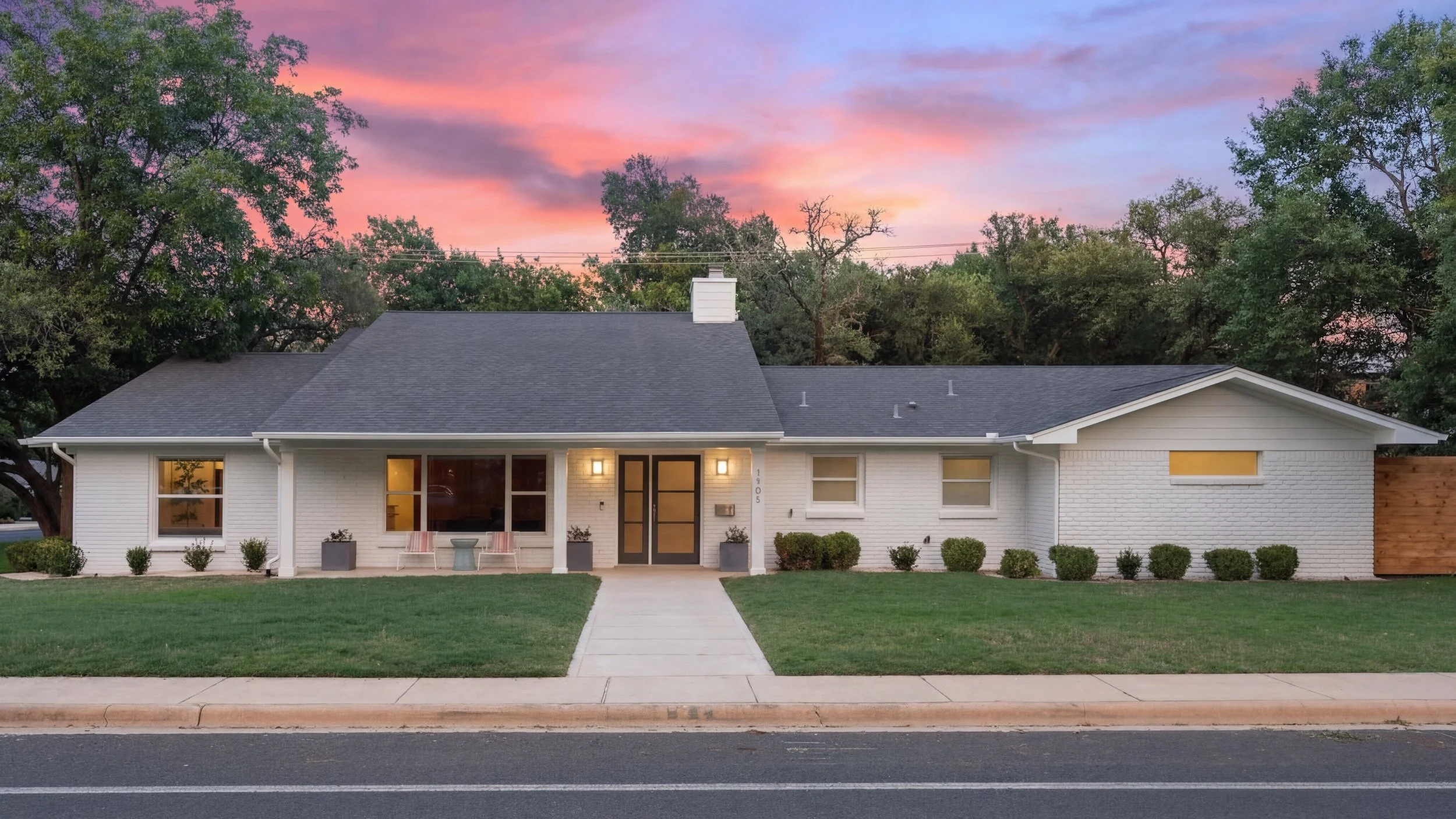A modern white brick house with a dark gray roof, front porch with two chairs, and a green lawn, during sunset sky with pink and purple clouds.