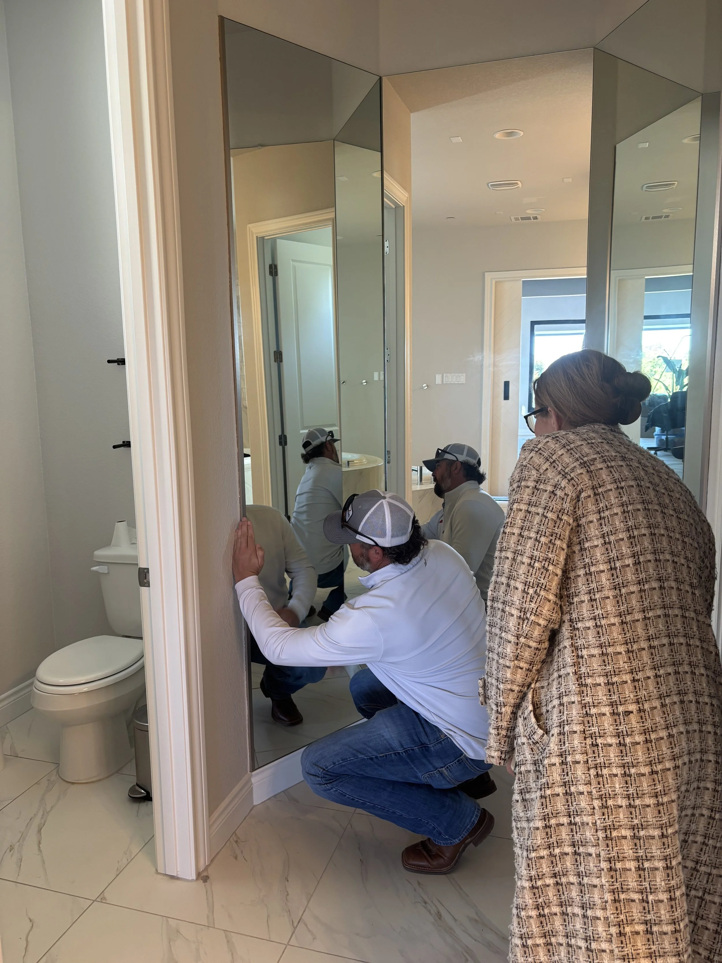 A woman observing a man squatting near a large wall mirror, with other people reflected in the mirror in a modern bathroom with white walls, beige marble flooring, a toilet, and a doorway leading to another room with large windows.