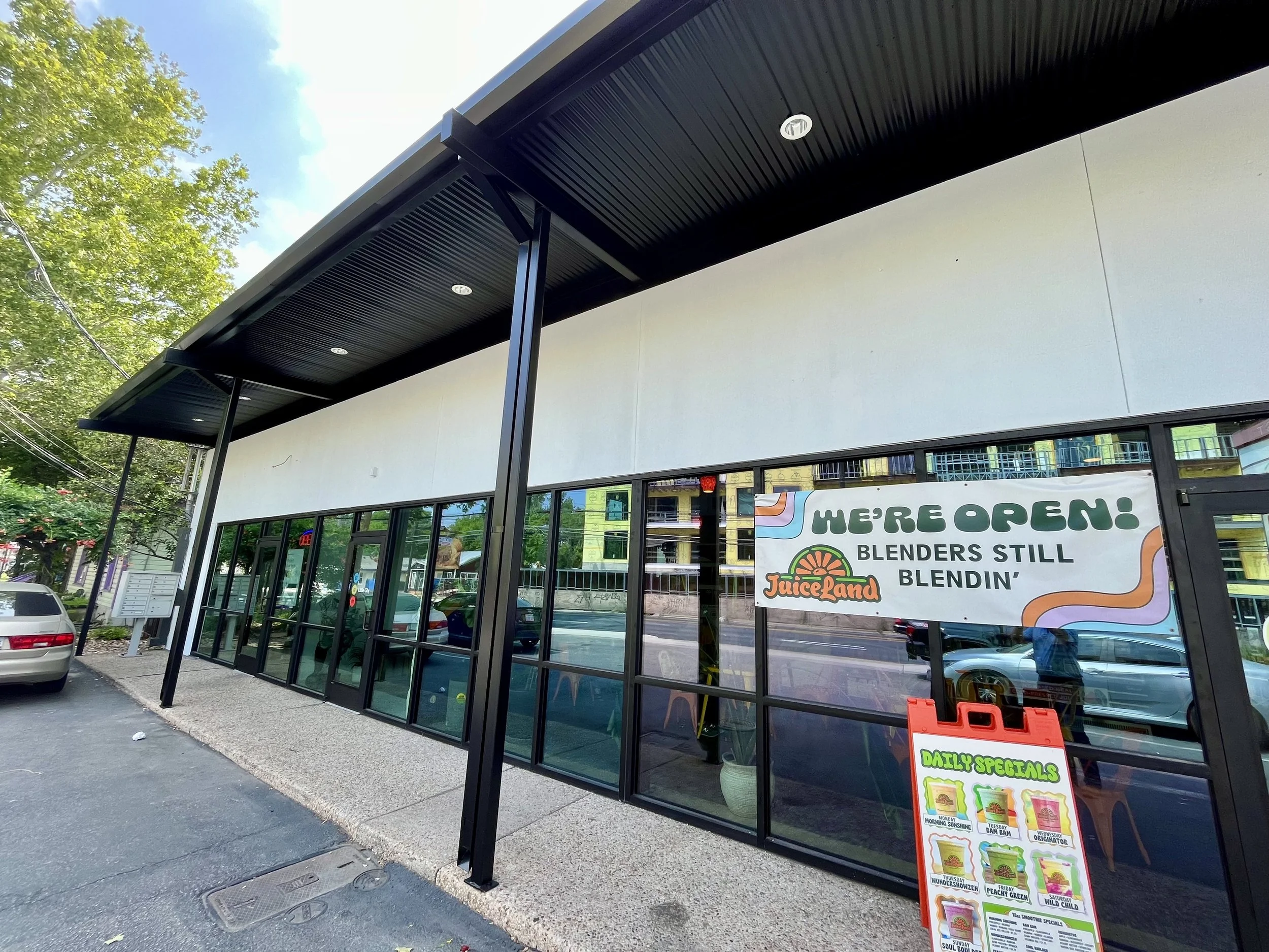 A storefront with large glass windows and hanging signs, including a banner that says 'WE'RE OPEN!' and 'BLENDERS STILL BLENDIN'' with the JuiceLand logo, an outdoor menu board displaying daily specials.
