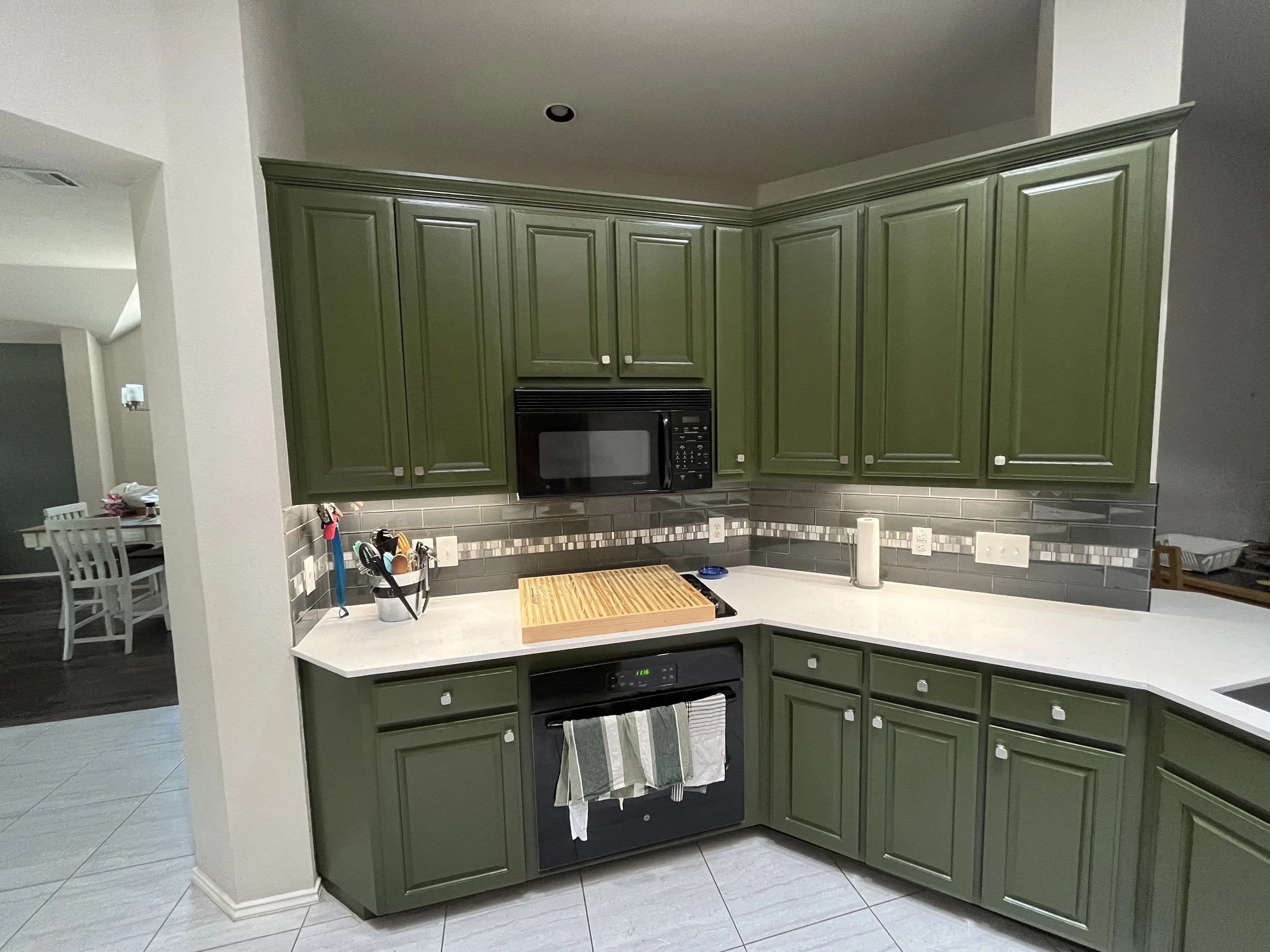 Green kitchen cabinets with black microwave oven above a white kitchen counter, tiled backsplash, paper towels, and various kitchen utensils, with a partial view of a dining area in the background.