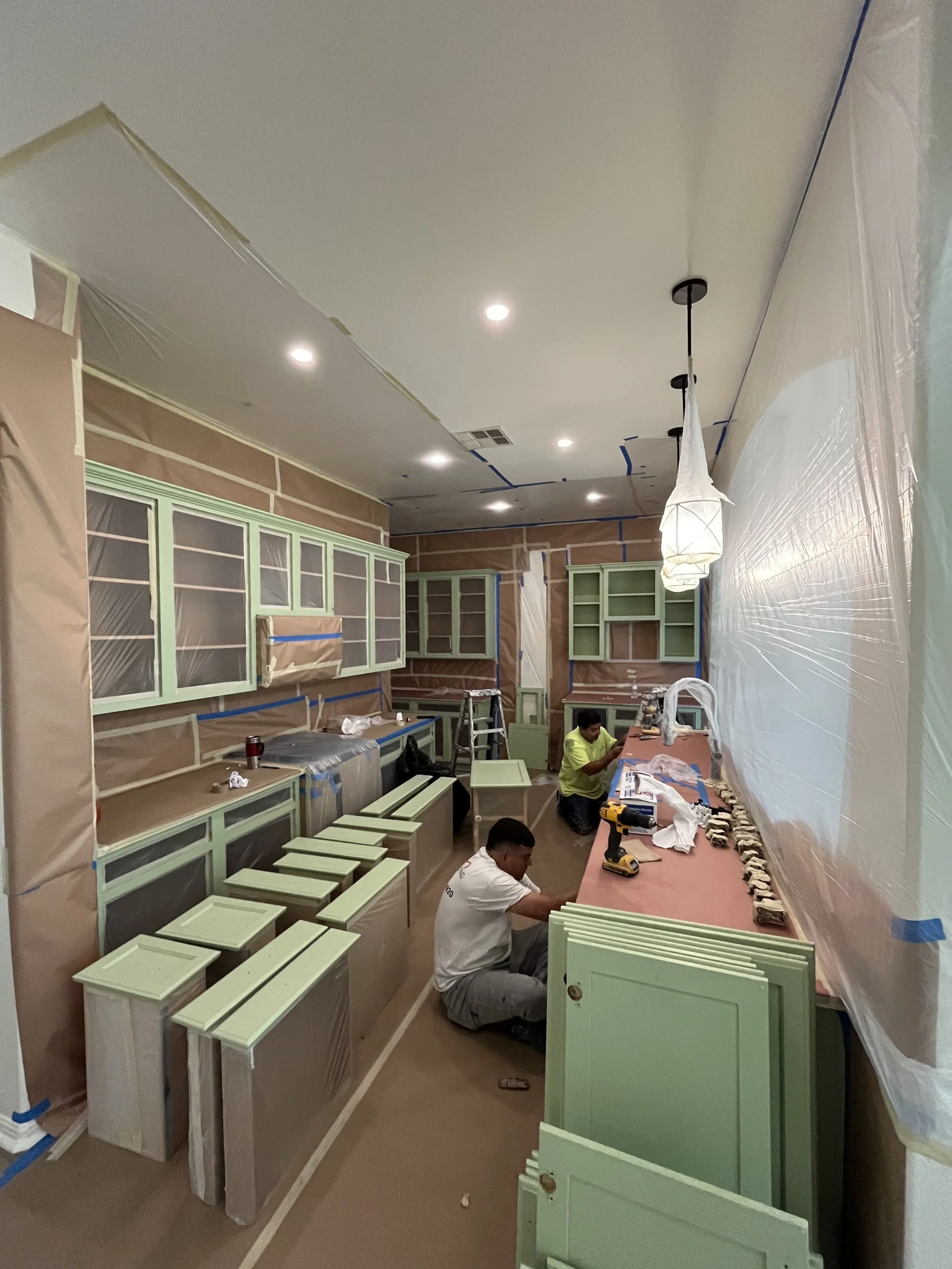 Kitchen renovation with light green cabinets, brown countertops, and ongoing painting or carpentry work. Two workers are installing or finishing cabinets and countertop area, surrounded by tools and supplies. Plastic covers and painter's tape protect