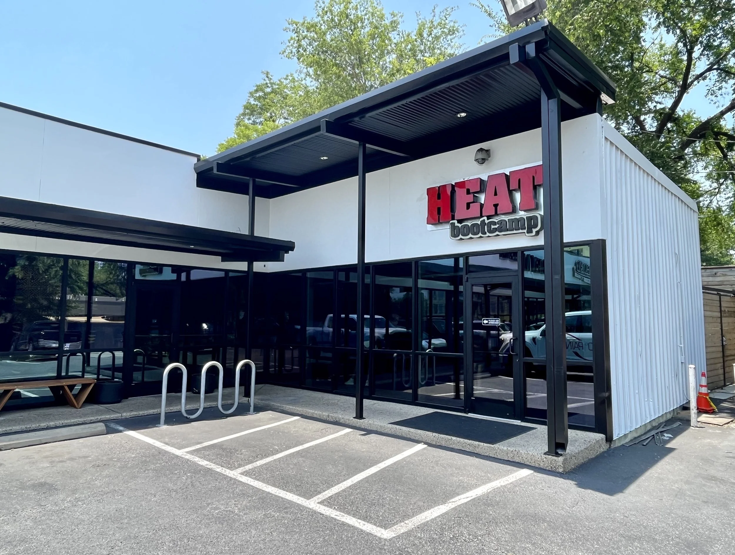 Exterior of a modern building with a sign that reads 'Heat Bootcamp', large windows, and a small parking lot with marked spaces in front. There is a covered seating area with a bench and bike racks. Trees are visible in the background under a blue sk