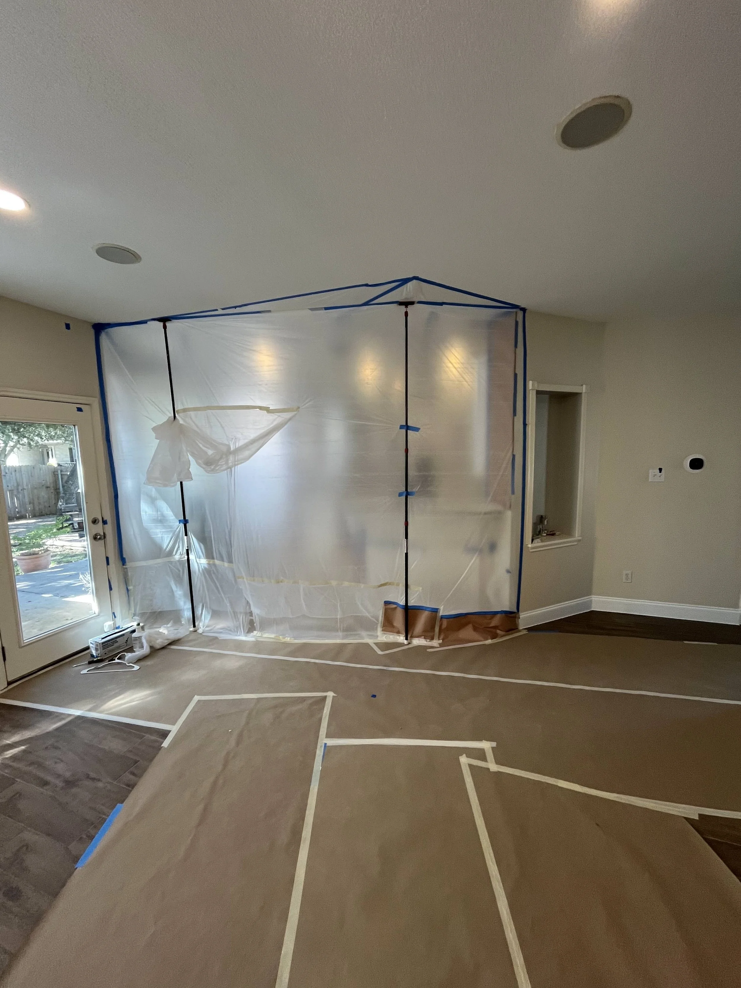 A room under renovation with painter's plastic sheeting set up to cover an area, and tape markings on the floor outlining designs for painting or carpeting