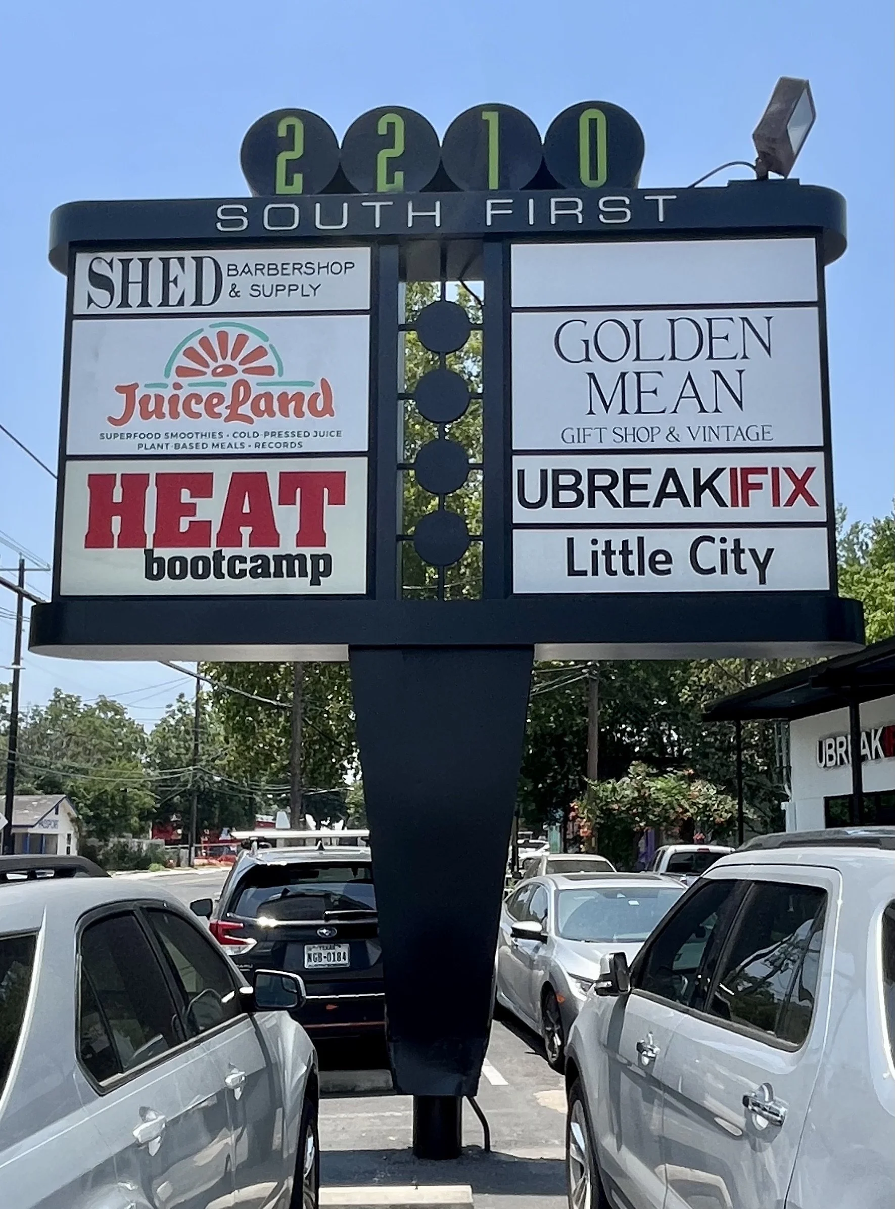 A parking lot with a digital sign displaying the date 2210 and the words 'South First'. There are parked cars and storefronts in the background, including a business called 'Ubreak iFix'.