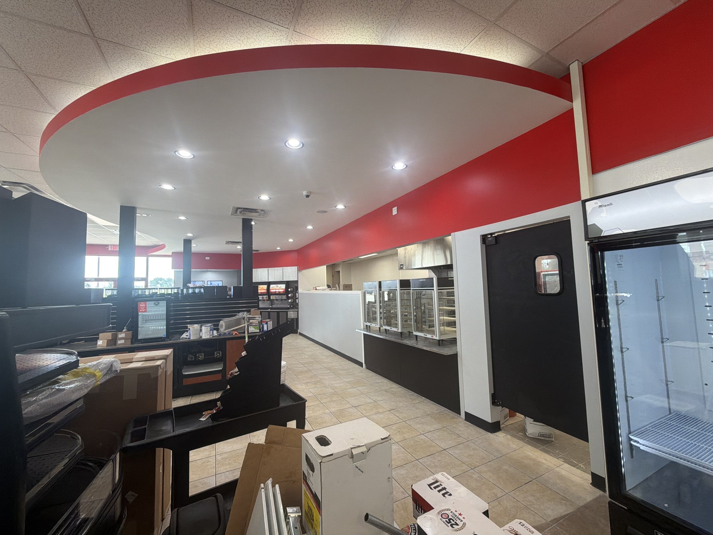 Interior of a store or restaurant under renovation or setup with boxes, shelves, and equipment scattered around, and a red and white ceiling.