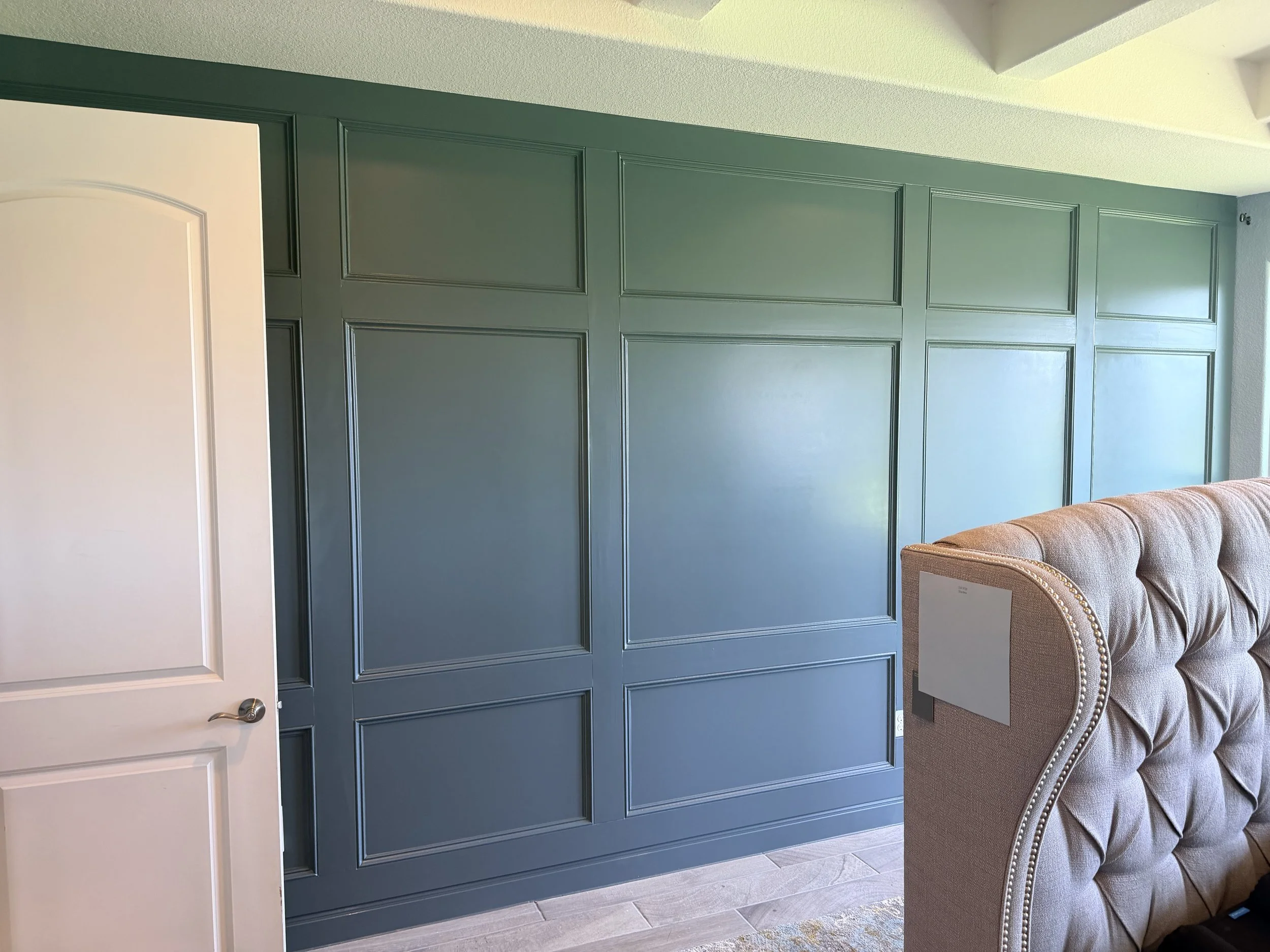 A room with teal-paneled wall, part of a white door with silver handle, and a beige tufted armchair with nailhead trim.