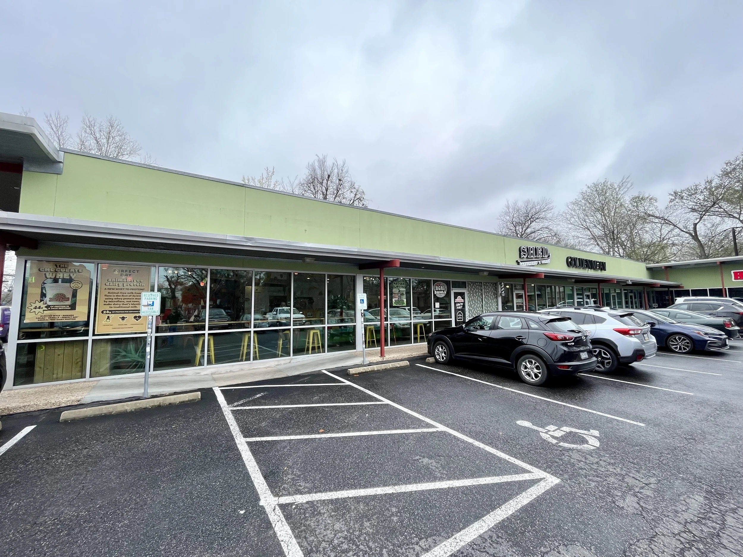 Parking lot with empty parking spaces in front of a shopping center. Cars are parked near the building with store signs visible. The sky is overcast.