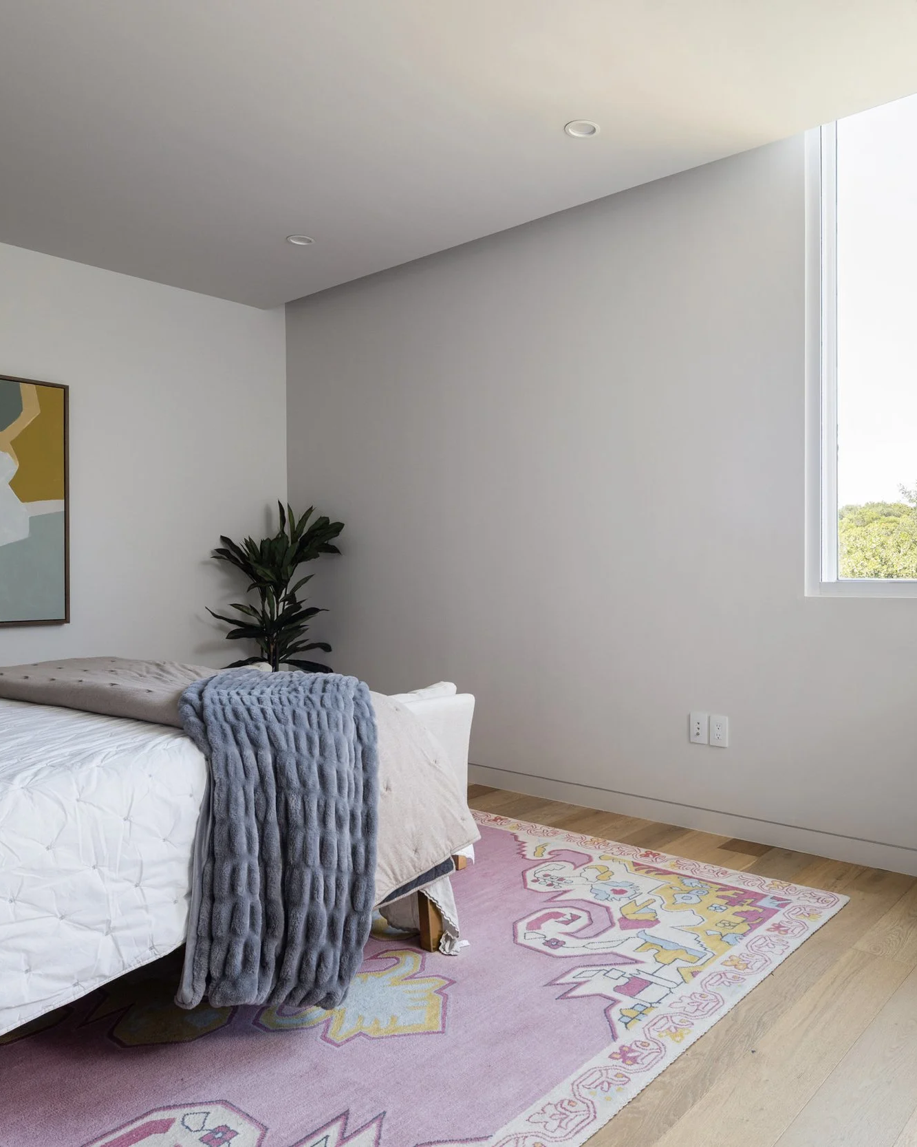 Part of a bedroom with a bed, a pink and yellow patterned rug, a green plant, a framed artwork, and a window showing trees outside.