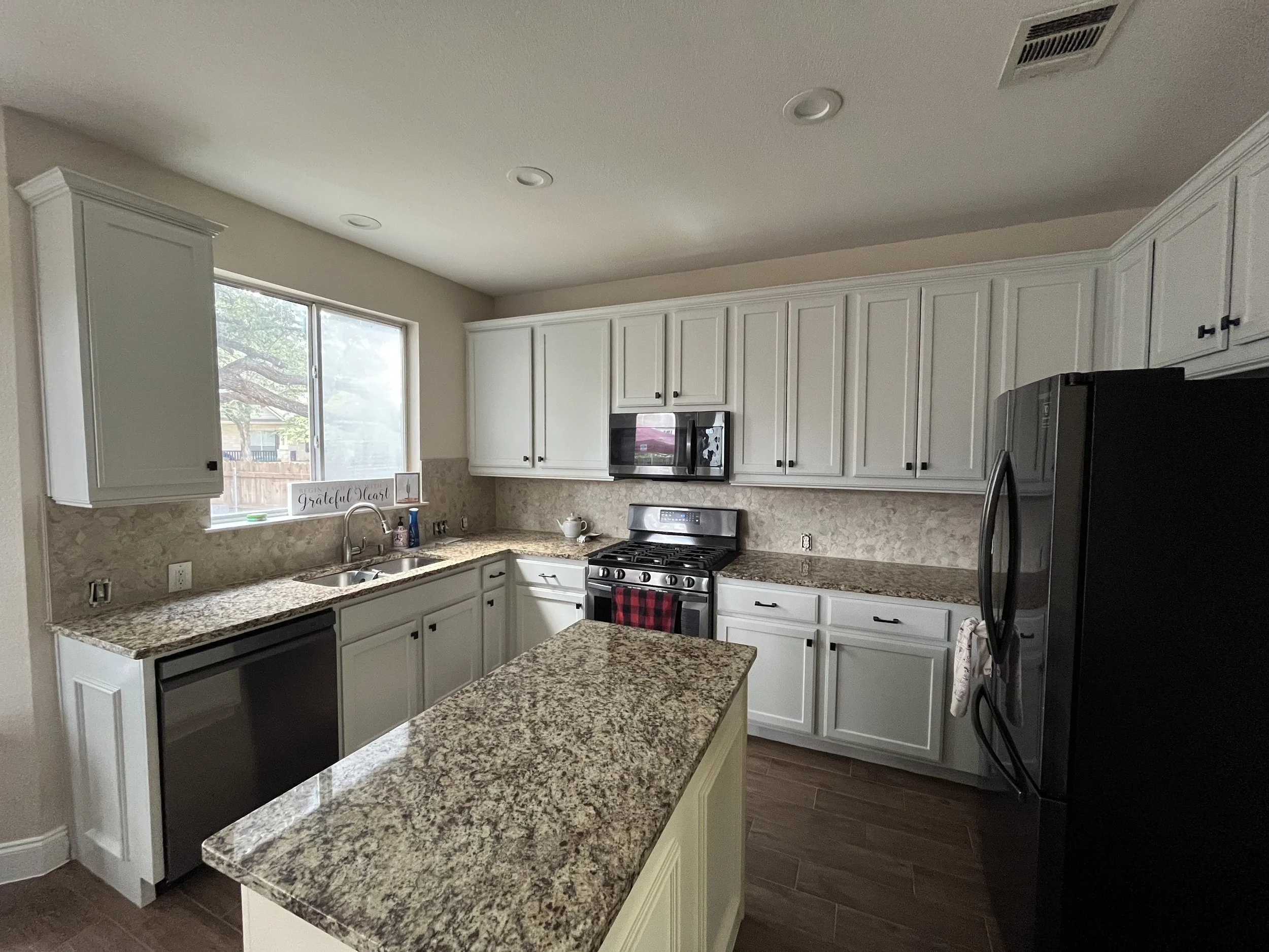 Kitchen with white cabinets, granite countertops, a window above the sink, stainless steel appliances including a microwave and a refrigerator, and a sign that reads "Grateful Heart" on the windowsill.