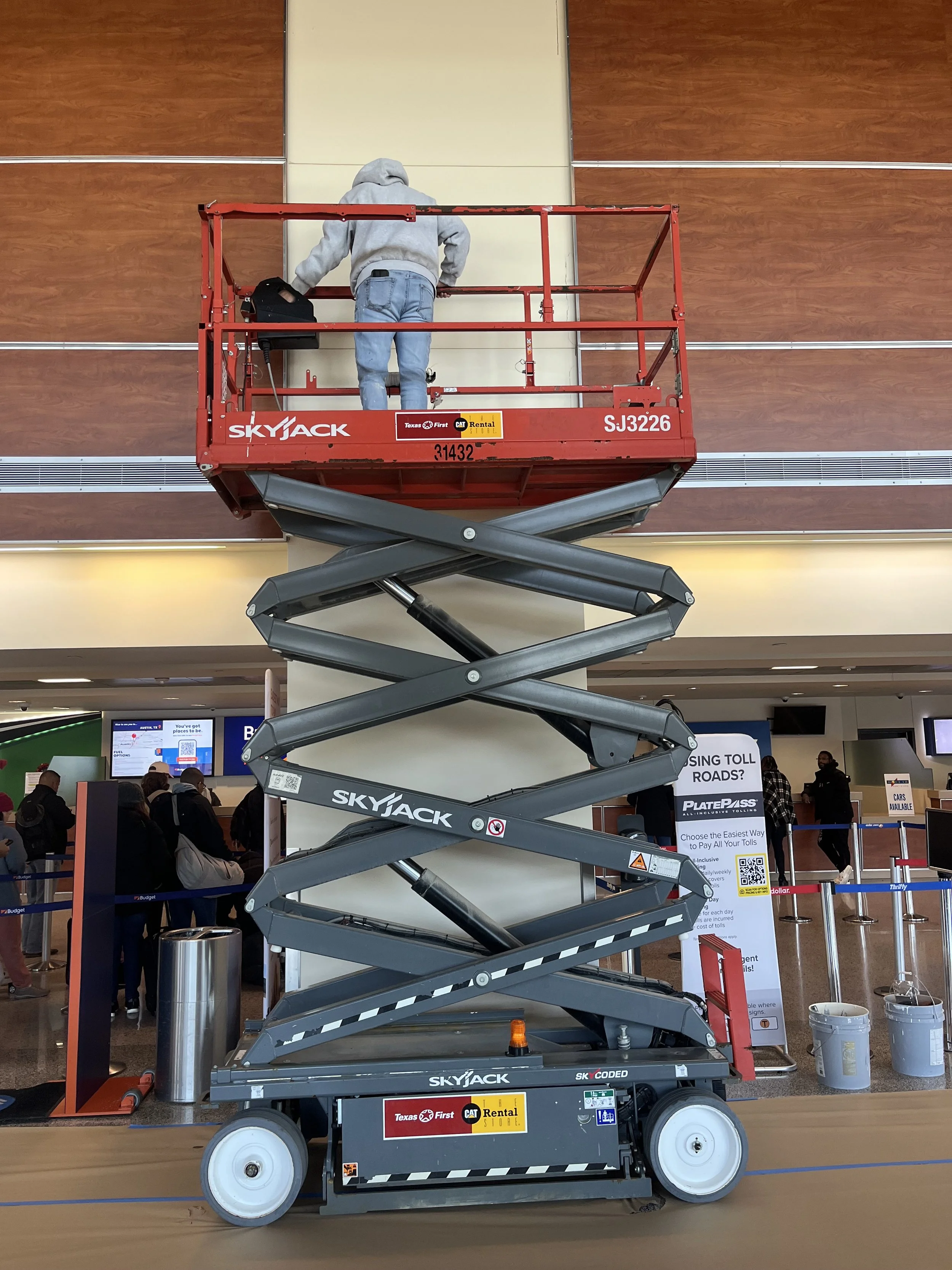 Airport terminal with a person on a red scissor lift near a wall, and luggage carts below. The scissor lift has labels for Skyjack and rental company.