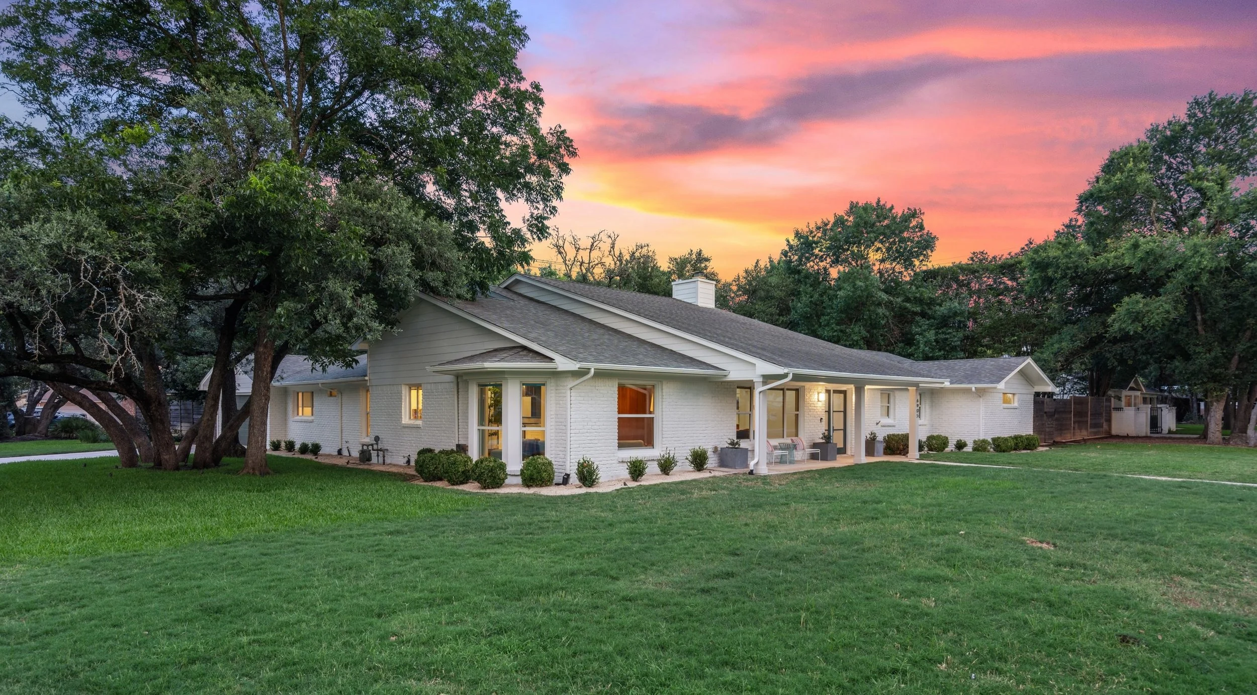A house repainted with Greek Villa from Sherwin-Williams with large windows and a covered porch, surrounded by green grass and trees, under a colorful sunset sky.