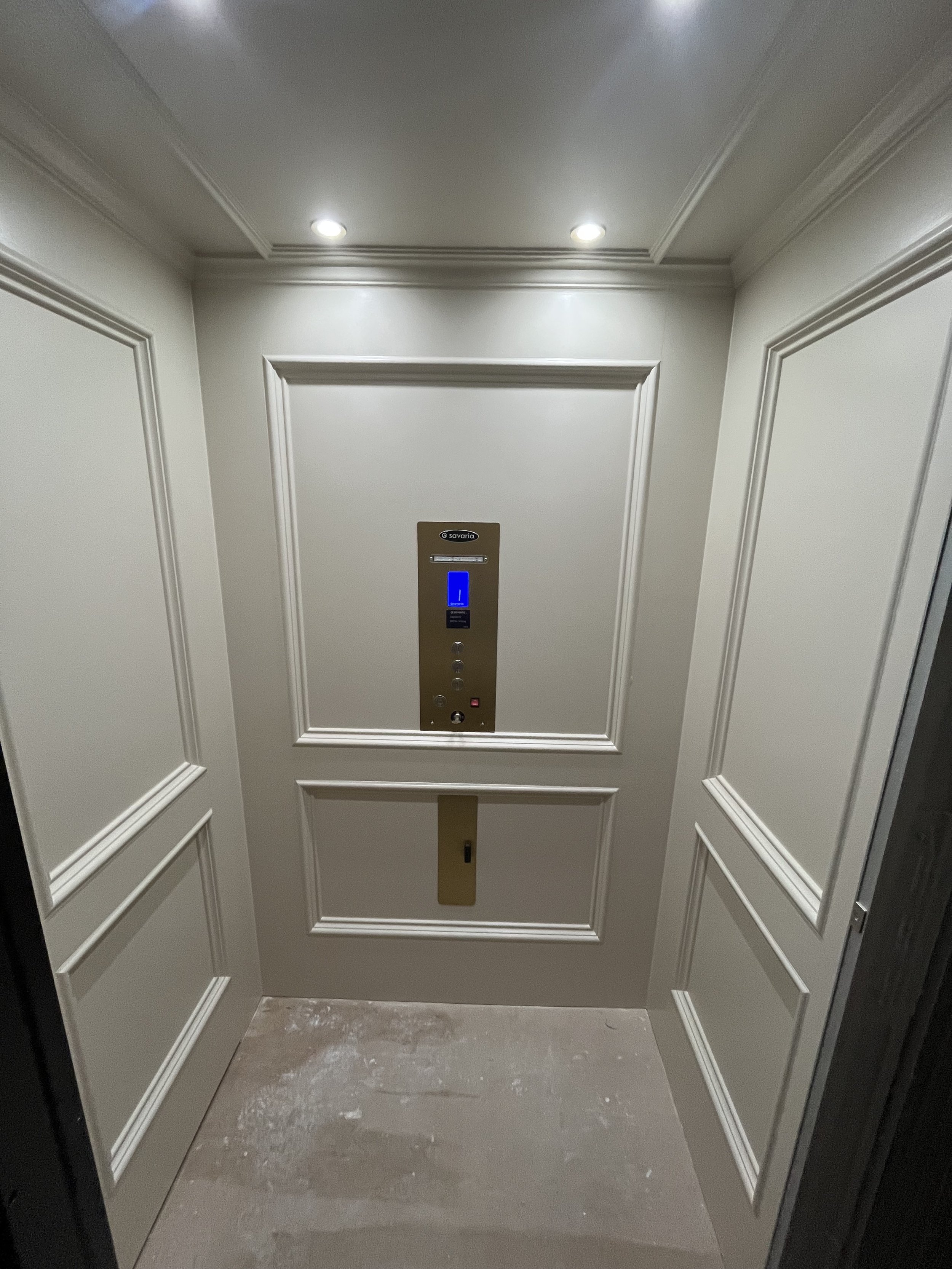 An elevator with closed doors, white panel walls with decorative molding, a control panel with digital display showing number 1, and a gray floor.