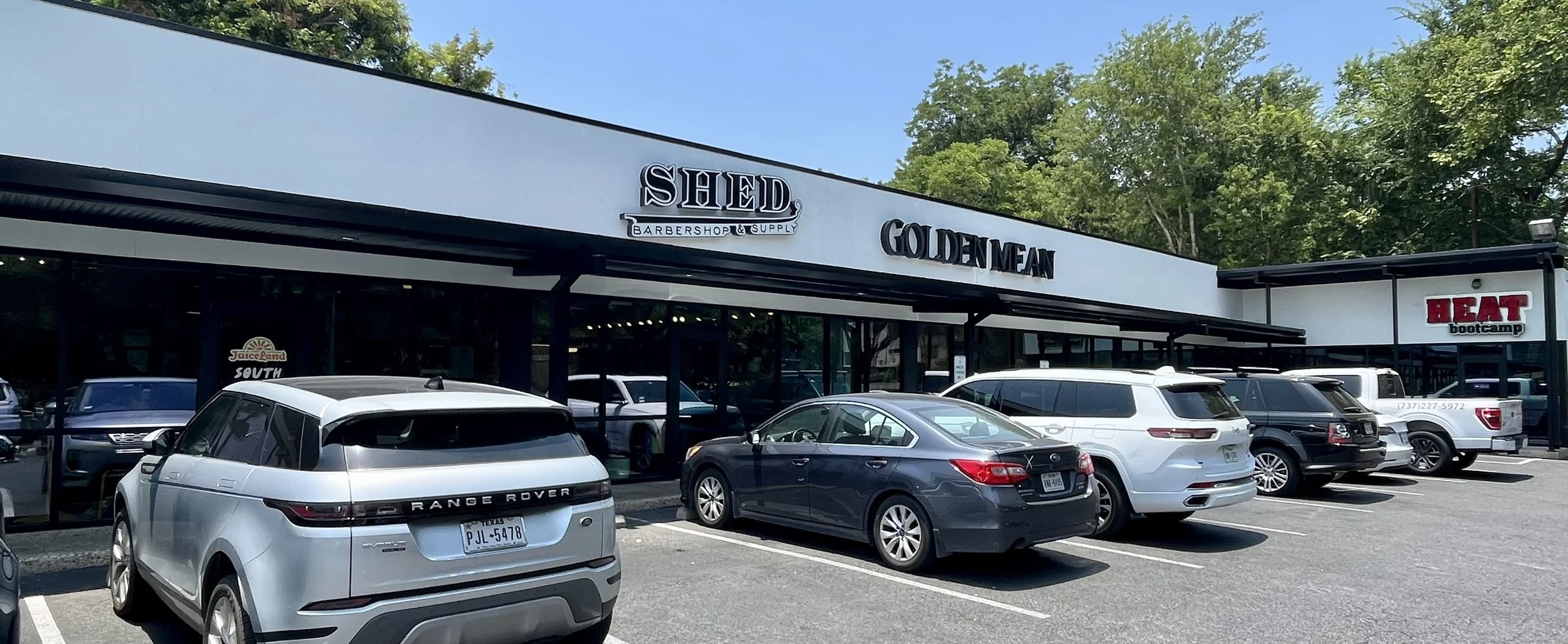 A parking lot in front of a strip mall with signs for Shed Barbershop & Supply, Golden Fan, and HEAT bootcamp. Several cars are parked, with trees and a blue sky in the background.