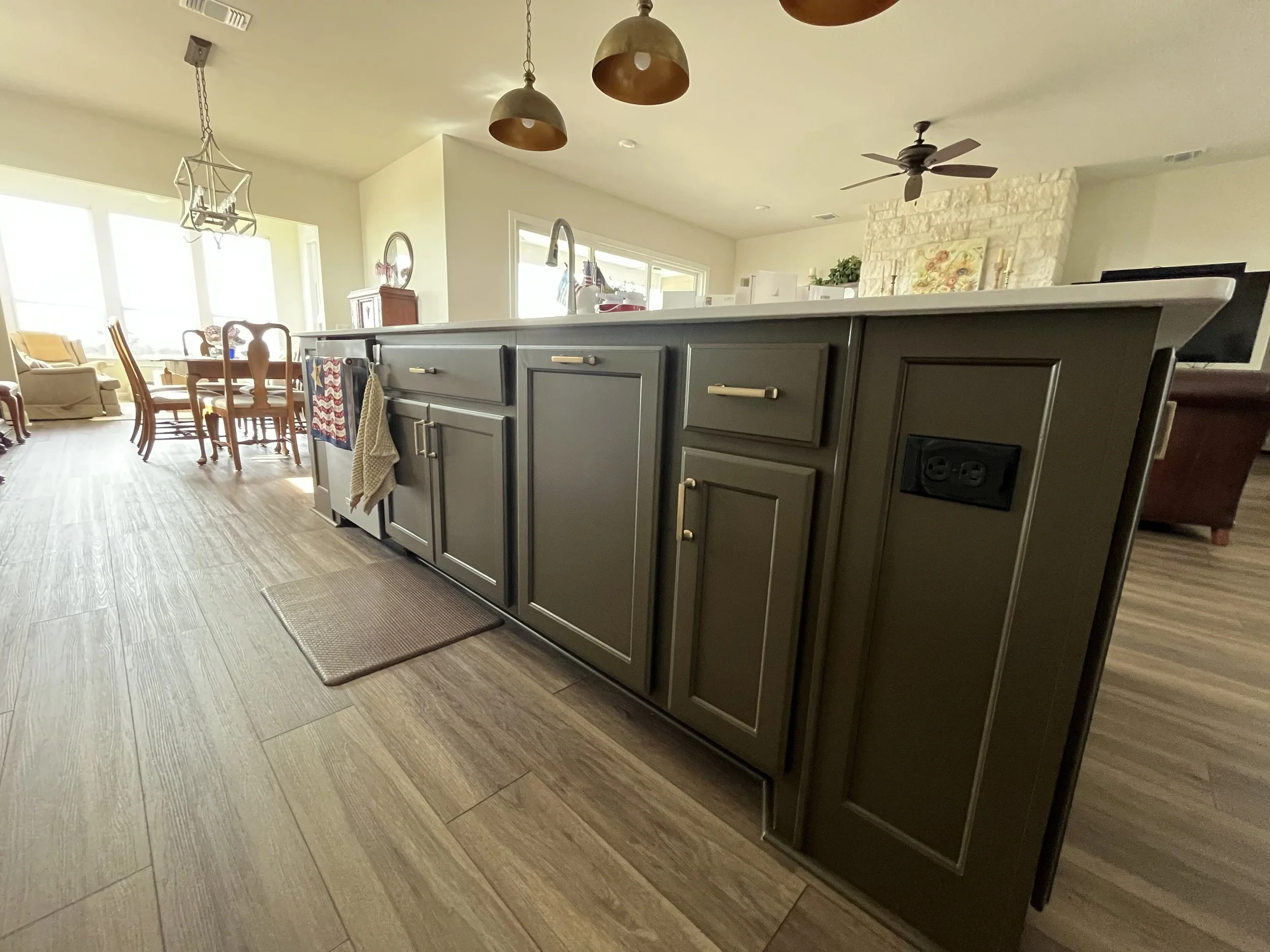 View of a kitchen island with dark gray cabinets, beige handles, and electrical outlets on the side, in a home with wood flooring, in front of a dining area and living room.