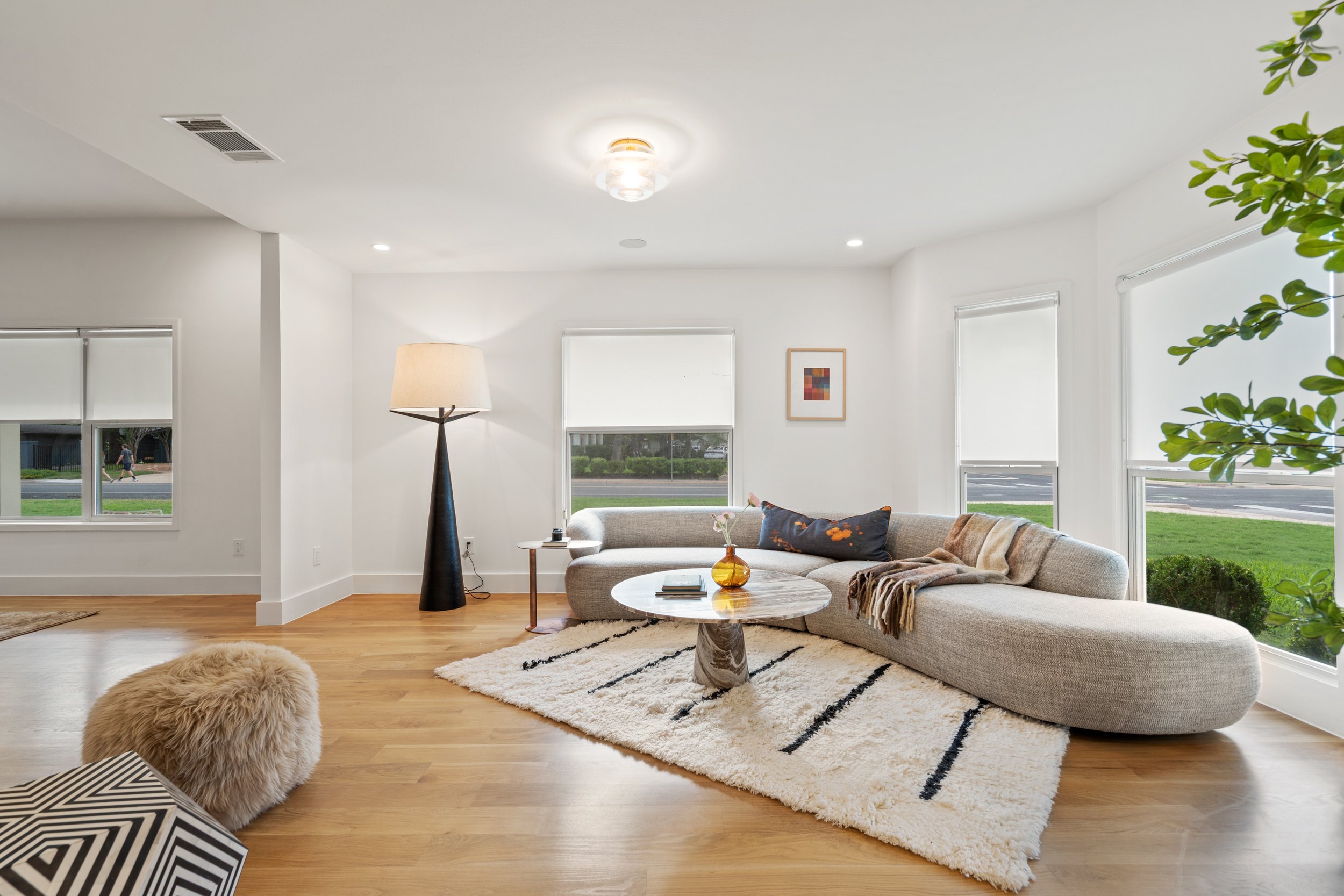 Bright living room with large windows, modern sofa, round marble coffee table, floor lamp, artwork, and greenery.