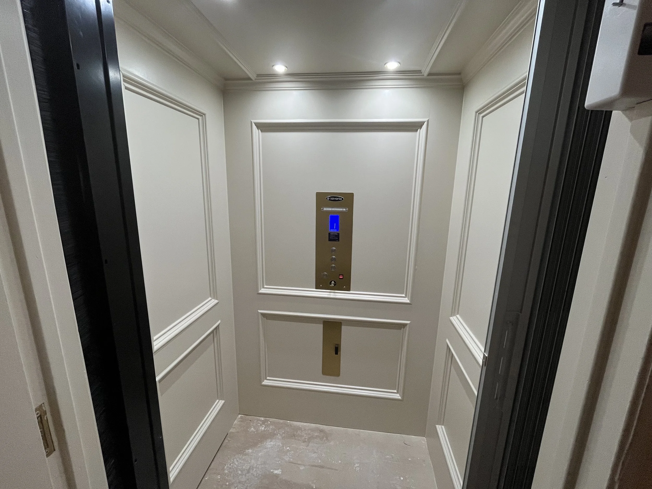 Inside an elevator with cream-colored paneled walls, ceiling with recessed lights, and a control panel with blue lights and buttons.