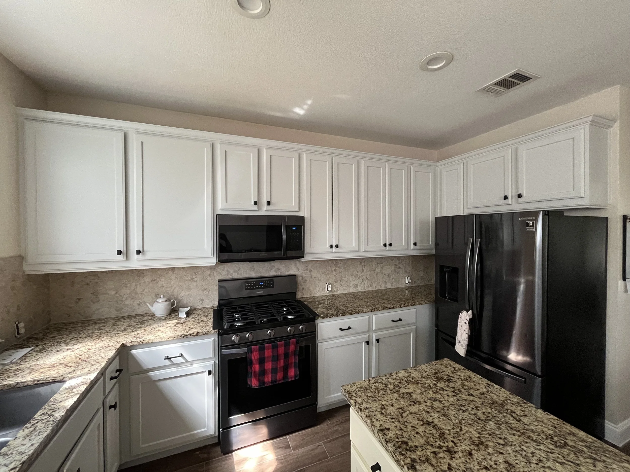 A kitchen with white cabinets, granite countertops, a stainless steel microwave and refrigerator, a stove with a red and black checkered dish towel, a tea set on the counter, and a tiled backsplash.