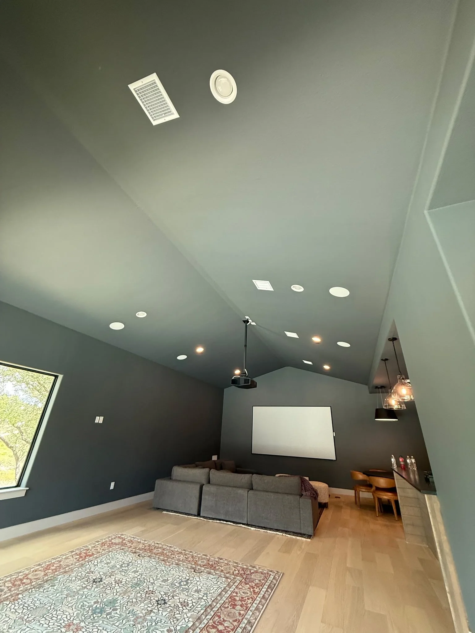 A modern living room with dark gray walls, a large gray sectional sofa, a white projector screen, a ceiling-mounted projector, and a window with natural light. There are hanging pendant lights and some water bottles on a countertop.