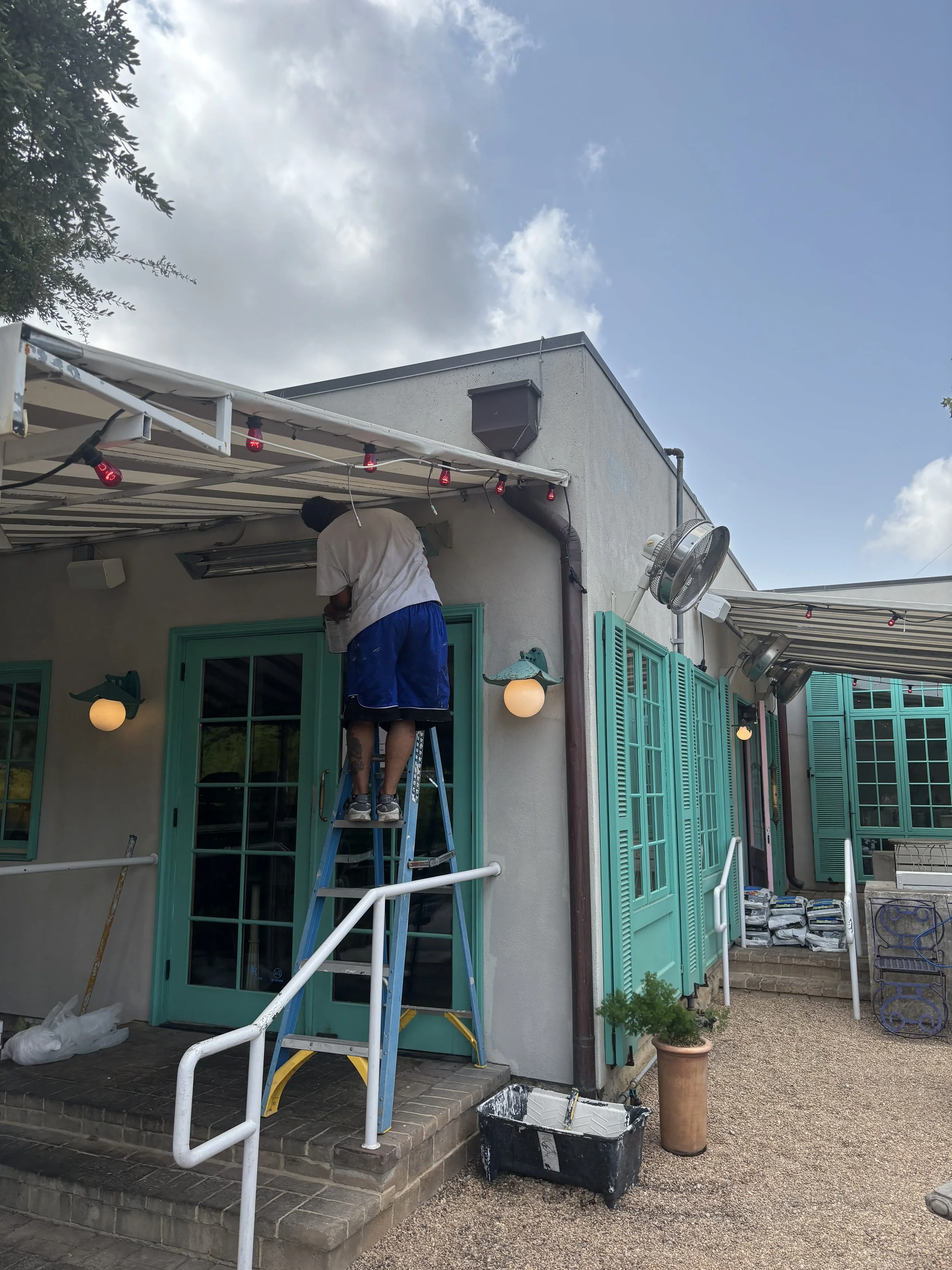 A person on a ladder working on the exterior of a house, with turquoise shutters and a door, under a blue sky with clouds, surrounded by potted plants and outdoor furniture.