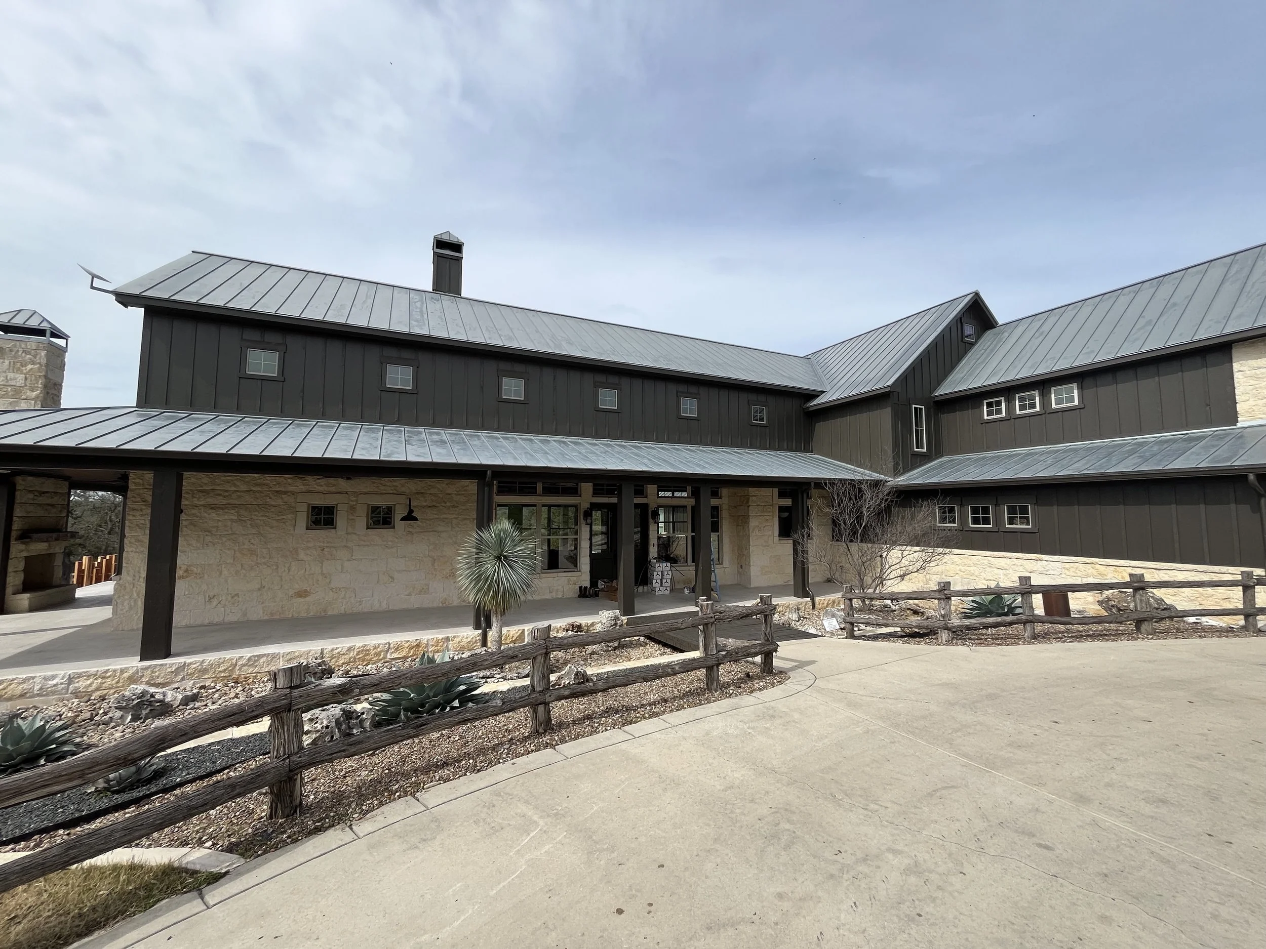 Modern house with black metal roof and beige stone walls, surrounded by a concrete driveway, landscaped with desert plants, and wooden fence, under a cloudy sky.
