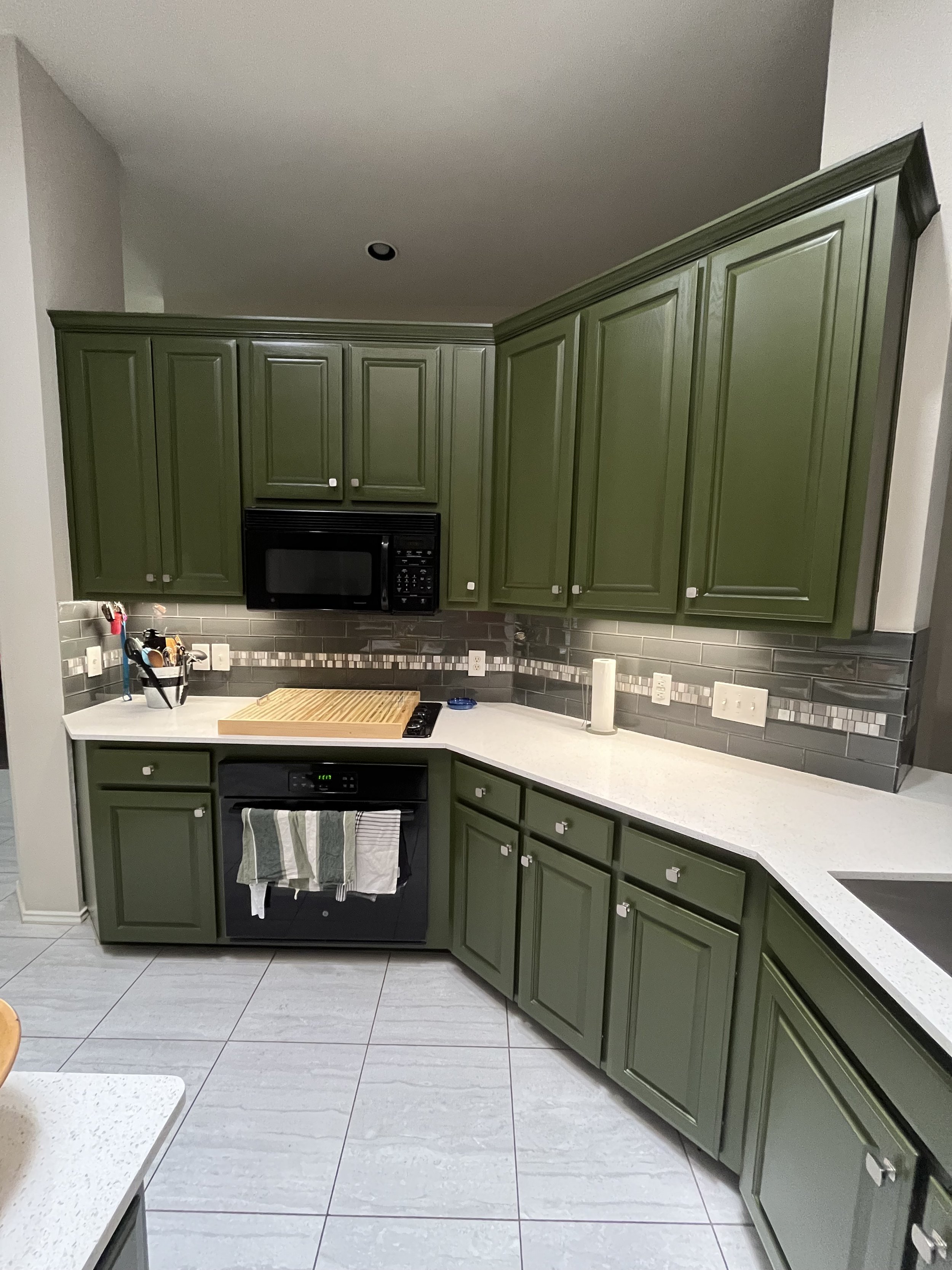 A kitchen with green cabinets, gray tile backsplash, white countertop, and various small appliances and utensils.