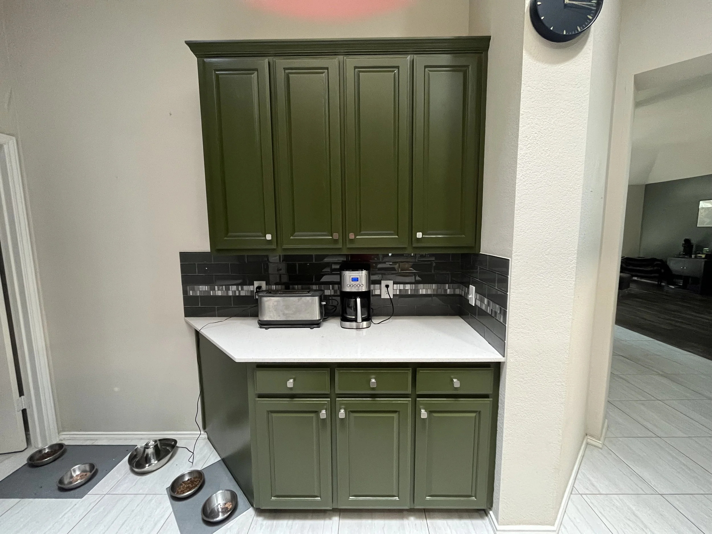 Green kitchen cabinet above a white countertop, with a toaster and coffee maker. Dog bowls on the floor and a black tiled backsplash.