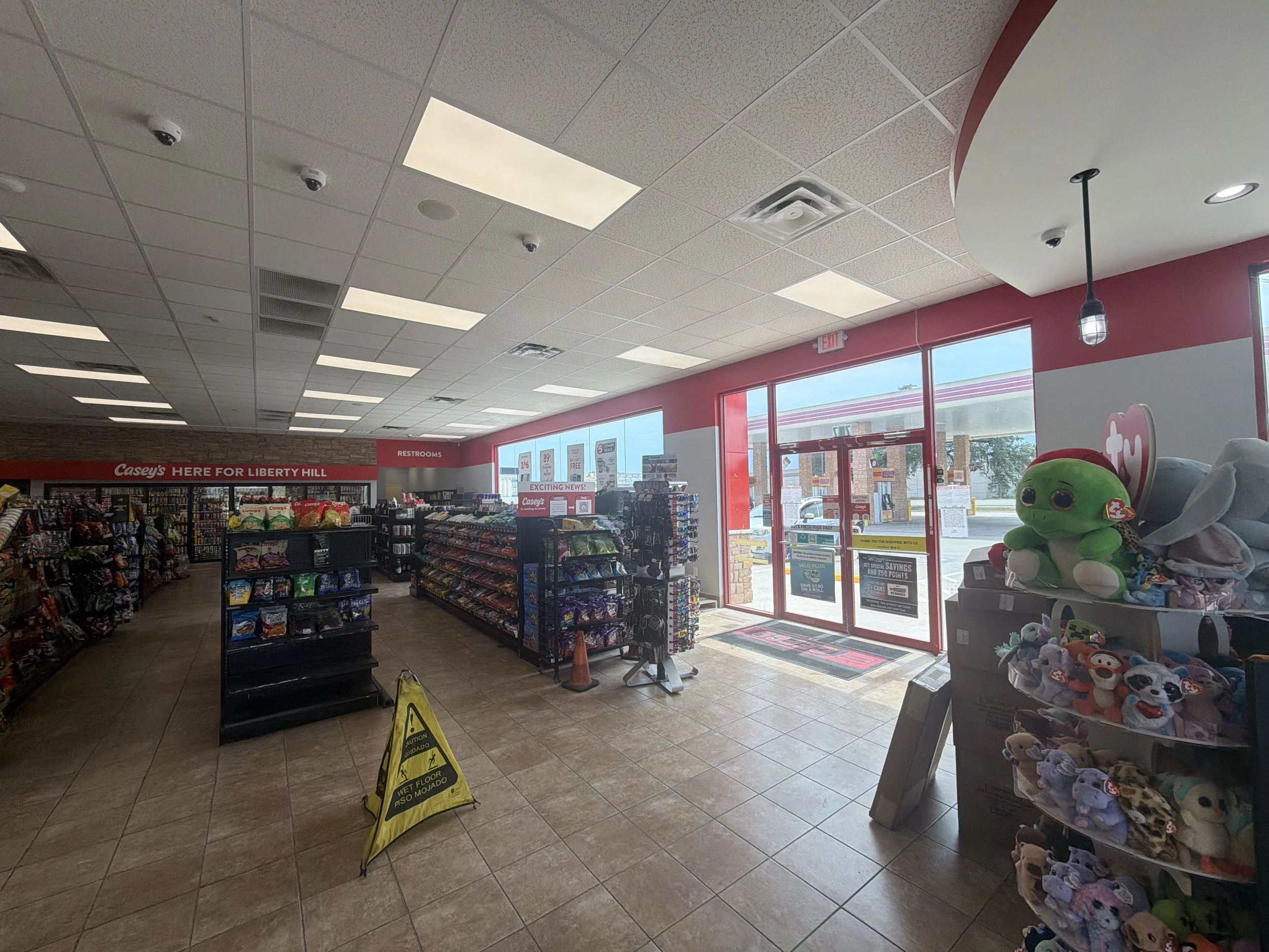 Inside a retail store with shelves of snacks, toys, and merchandise near the entrance with large glass windows and red trim. A yellow caution sign on the tiled floor indicates a wet floor.