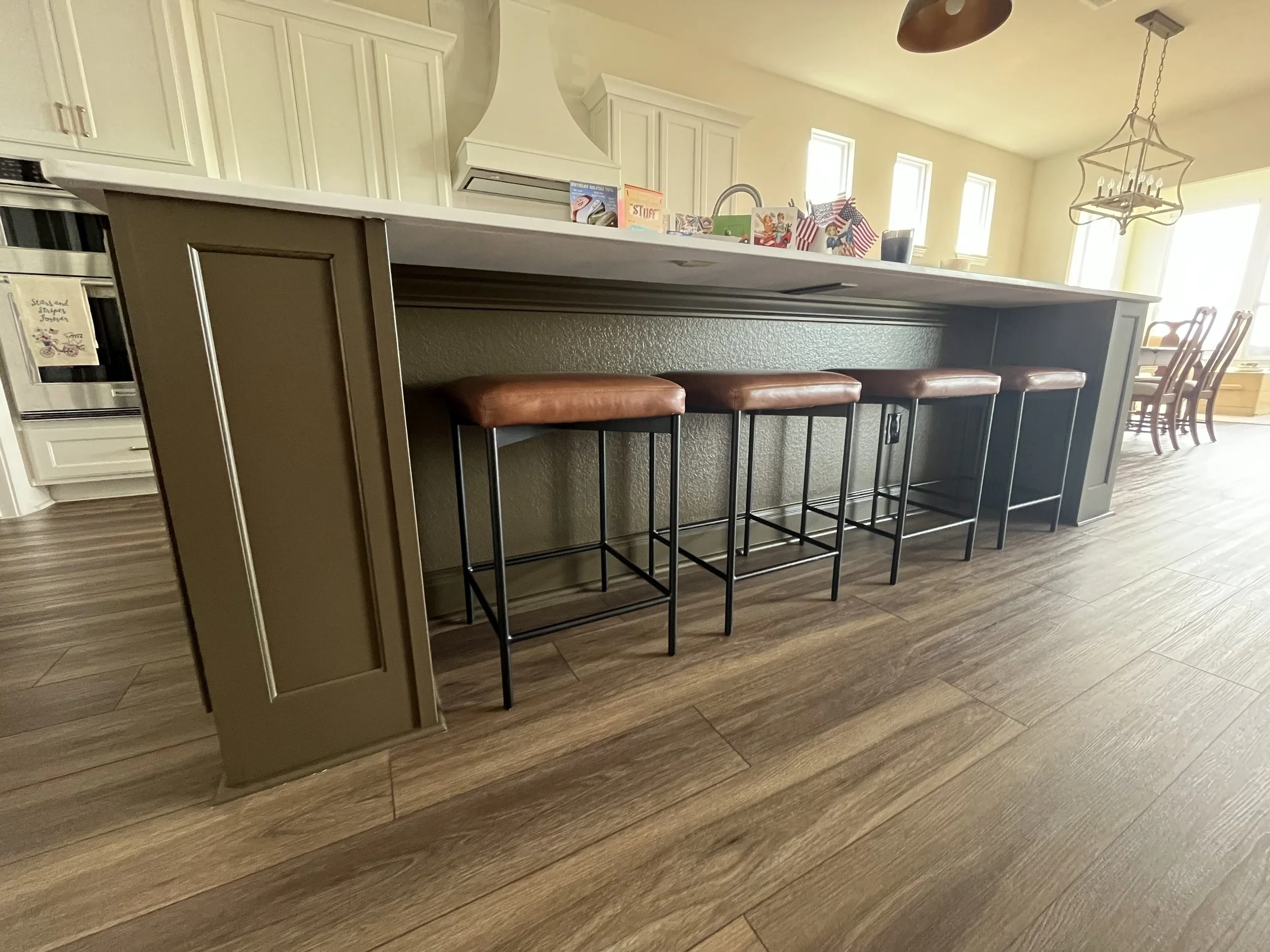 View of a kitchen island with four brown leather stools, featuring black metal legs, in a room with wooden flooring, white cabinets, and a dining area with chairs and large windows in the background.