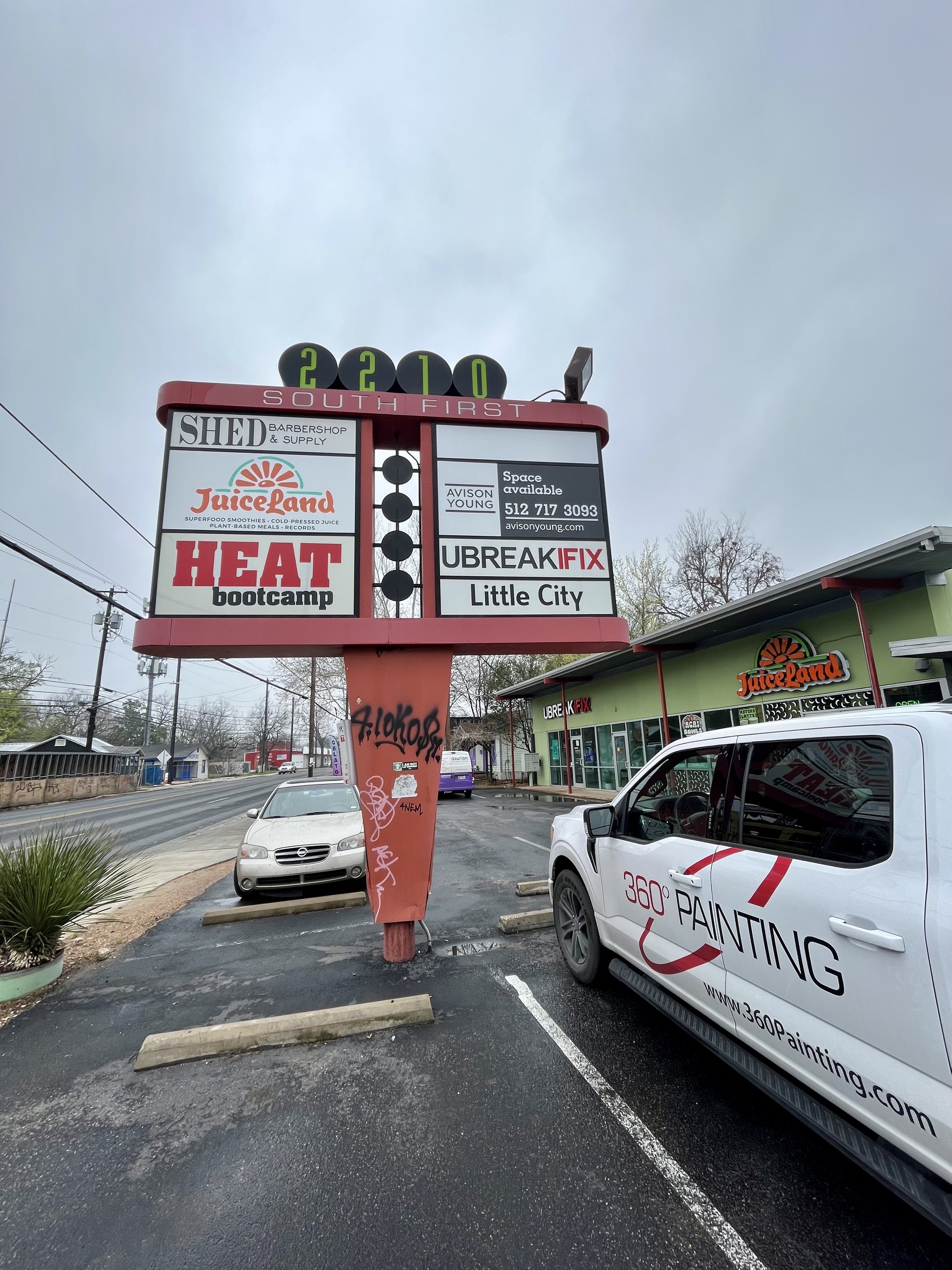 A parking lot outside a strip mall with a sign displaying businesses such as JuiceLand, Heat Bootcamp, and UBreakiFix. There are vehicles parked, including a white van with a painted logo for 360 Painting. The sky is overcast.