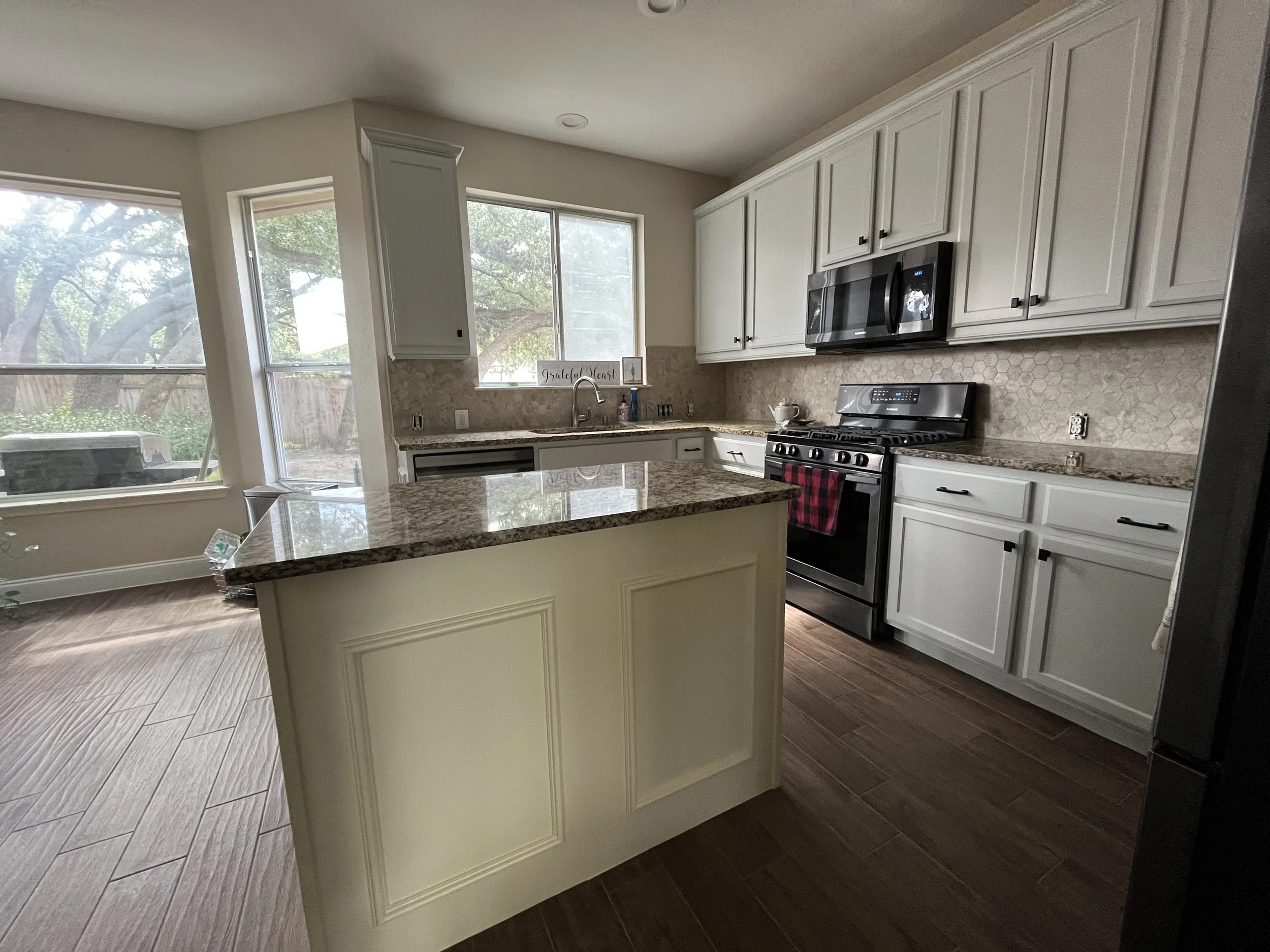 A kitchen with white cabinets, black appliances, a granite countertop, and large windows showing trees outside.