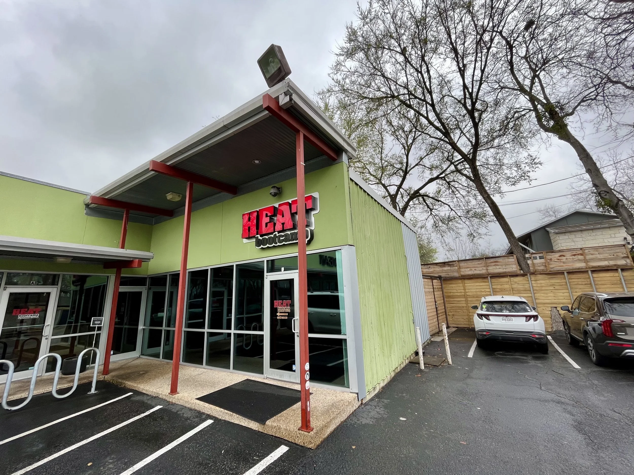 Exterior of a Green building with a red and white sign that reads 'Heat bootcamp.' The building has a glass entrance door, metal bike racks, and parking lot with white painted lines. There are cars parked and a wooden fence in the background. A leafl