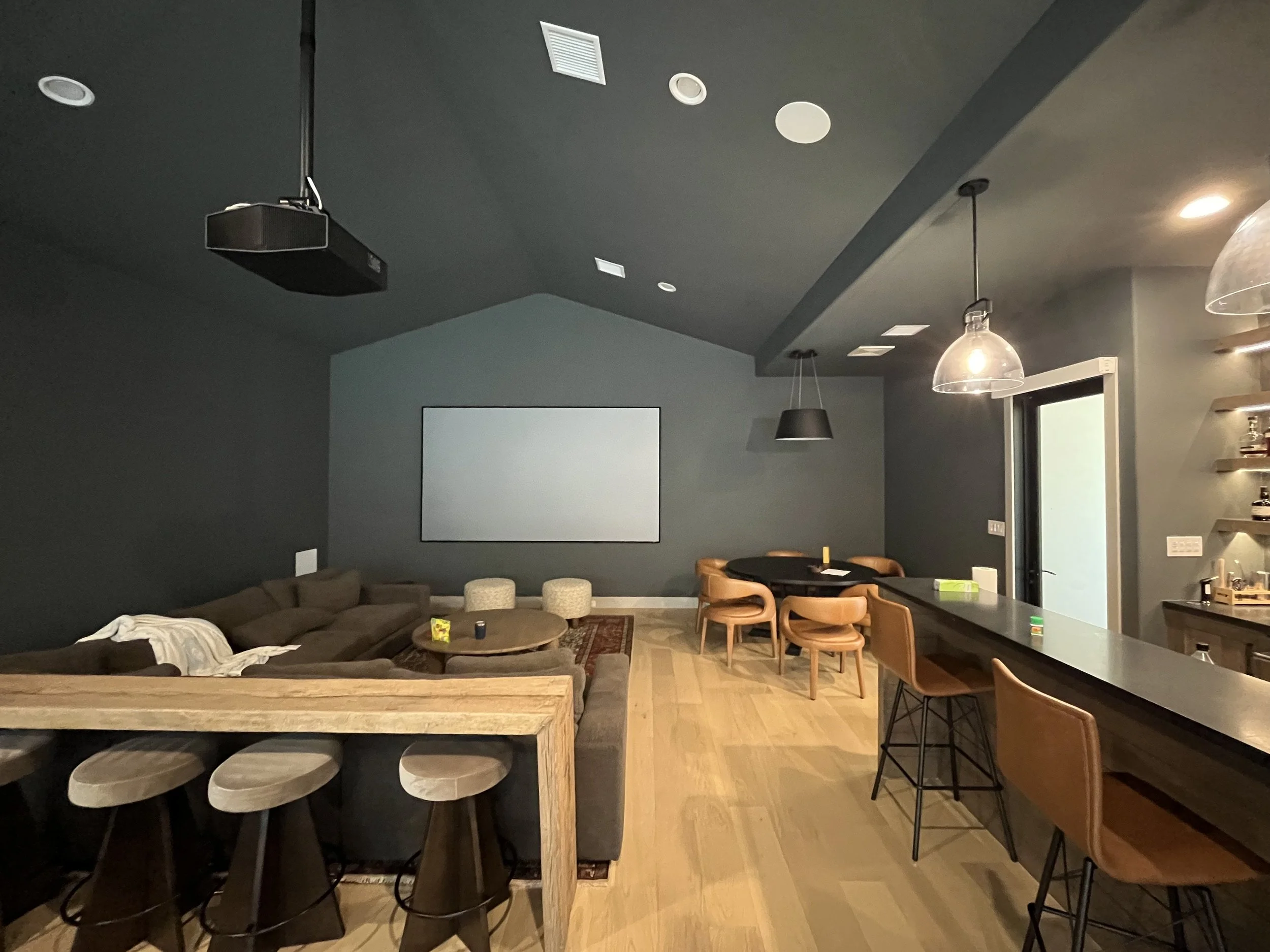 Interior of a modern living room with a dark ceiling, dark gray walls, a brown sectional sofa with pillows, a wooden counter with stools, a round dining table with chairs, hanging light fixtures, and a window with a sliding door.