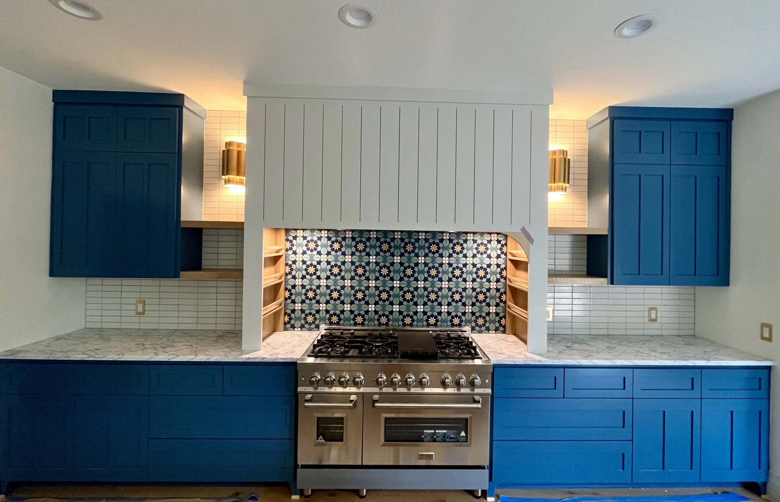 Modern kitchen with blue cabinets, marble countertops, a stainless steel oven with a gas stove, and a decorative tiled backsplash behind the stove.