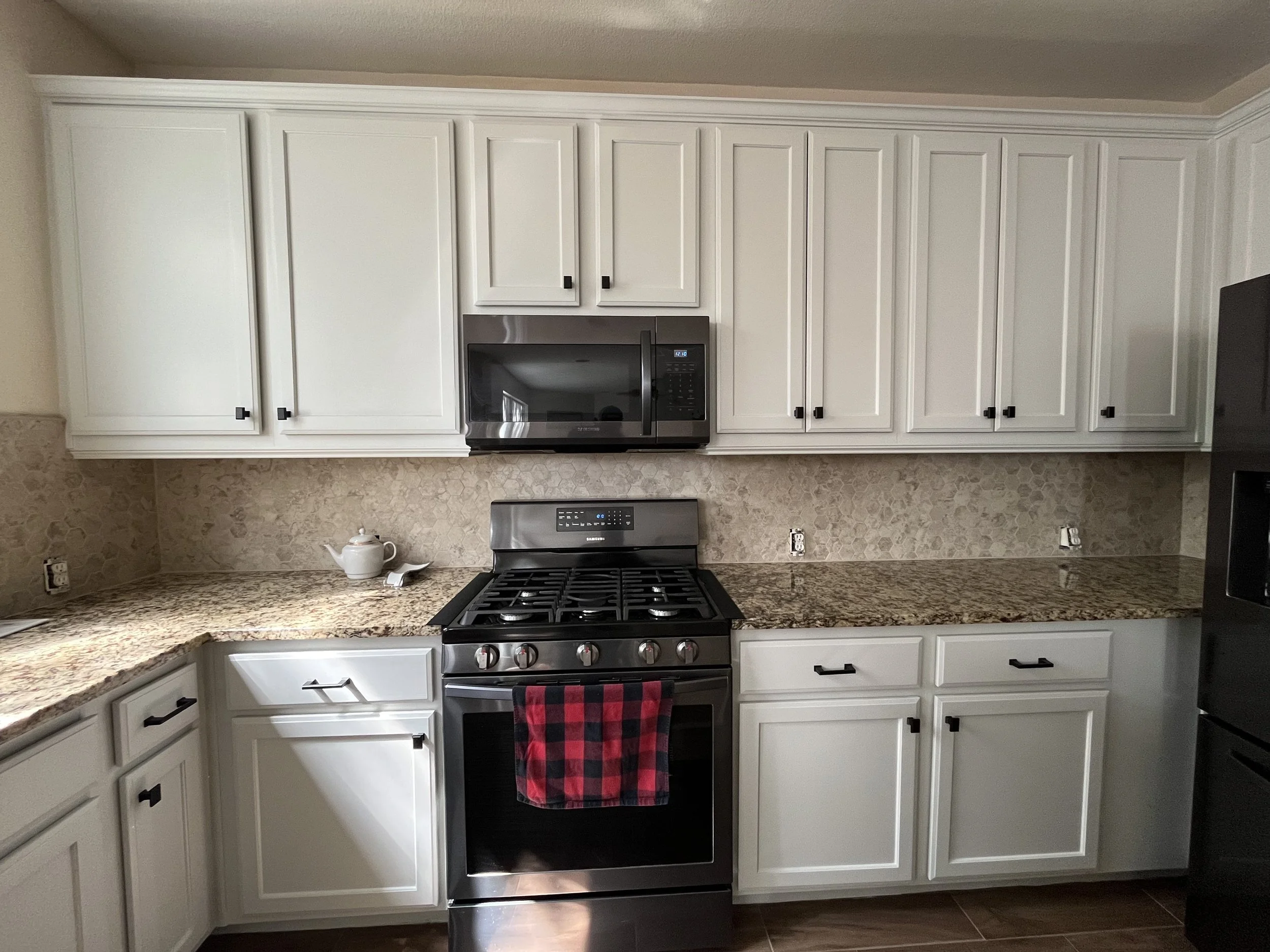 White kitchen cabinets with black handles, stainless steel microwave above stove, granite countertops, teapot, red and black checkered towel, and beige tile backsplash.