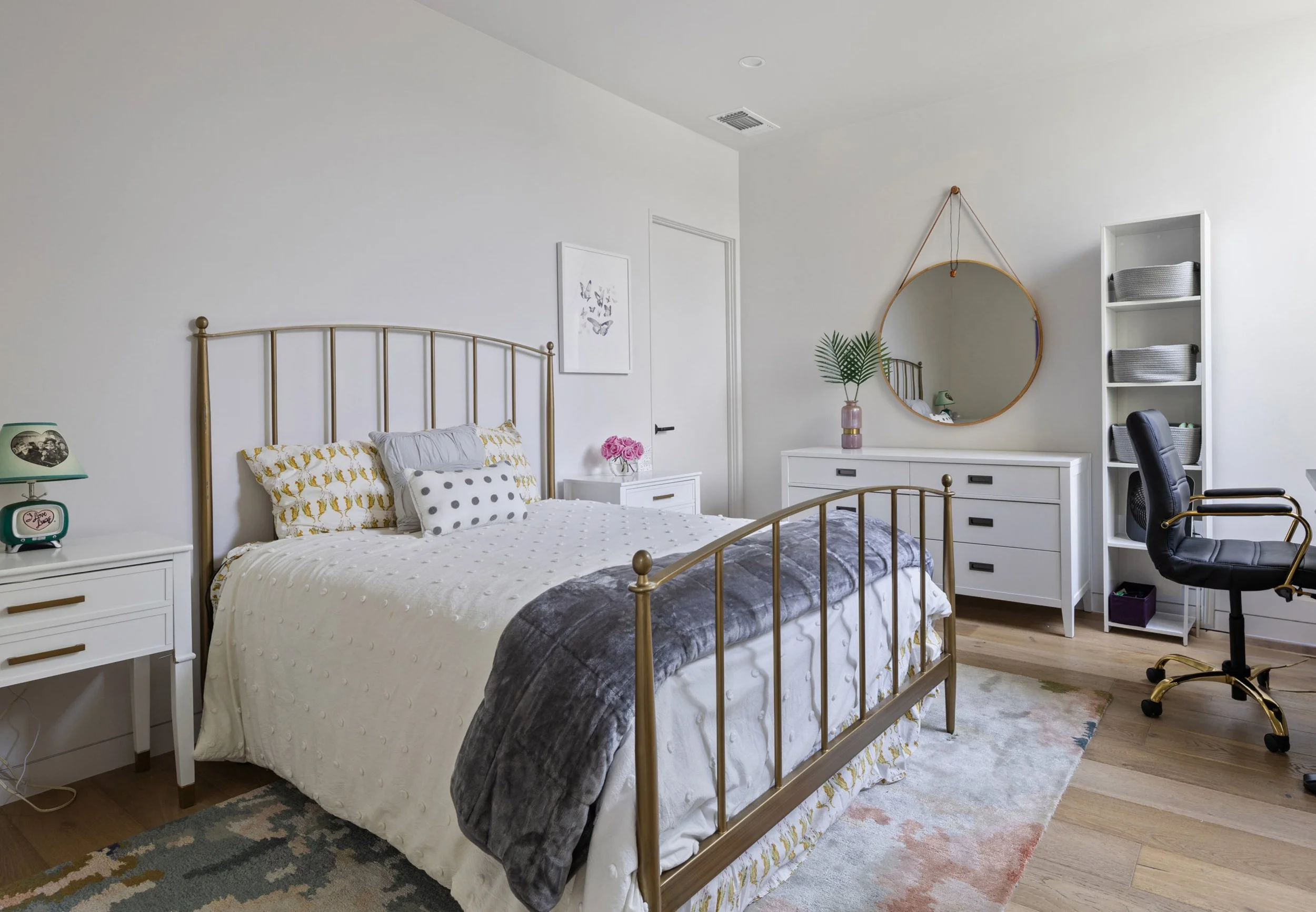 A bedroom with white walls, a large bed with a white dotted comforter and patterned pillows, gold metal bed frame, white nightstands with decor, a white dresser with a mirror and plant, a black office chair, a white shelving unit, and a colorful rug.