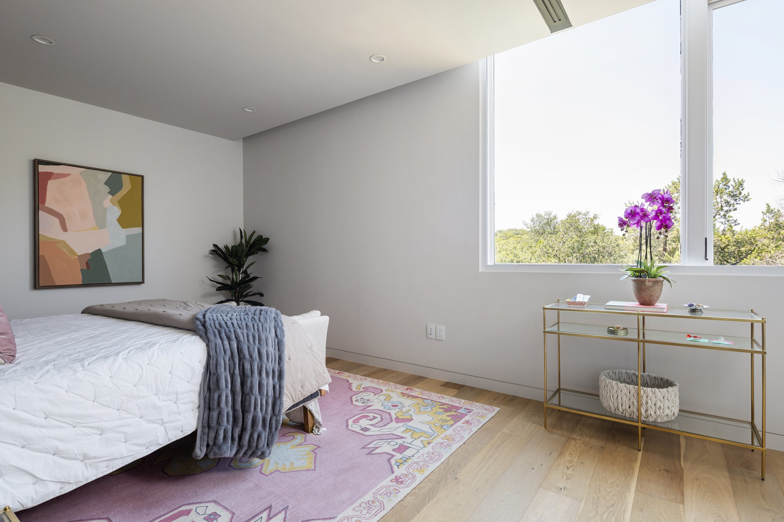 A bright bedroom with a bed covered in white bedding and a gray blanket, a colorful abstract painting on the wall, a potted plant in the corner, a window with a view of trees, and a gold-framed glass table with a pink orchid and decorative items.