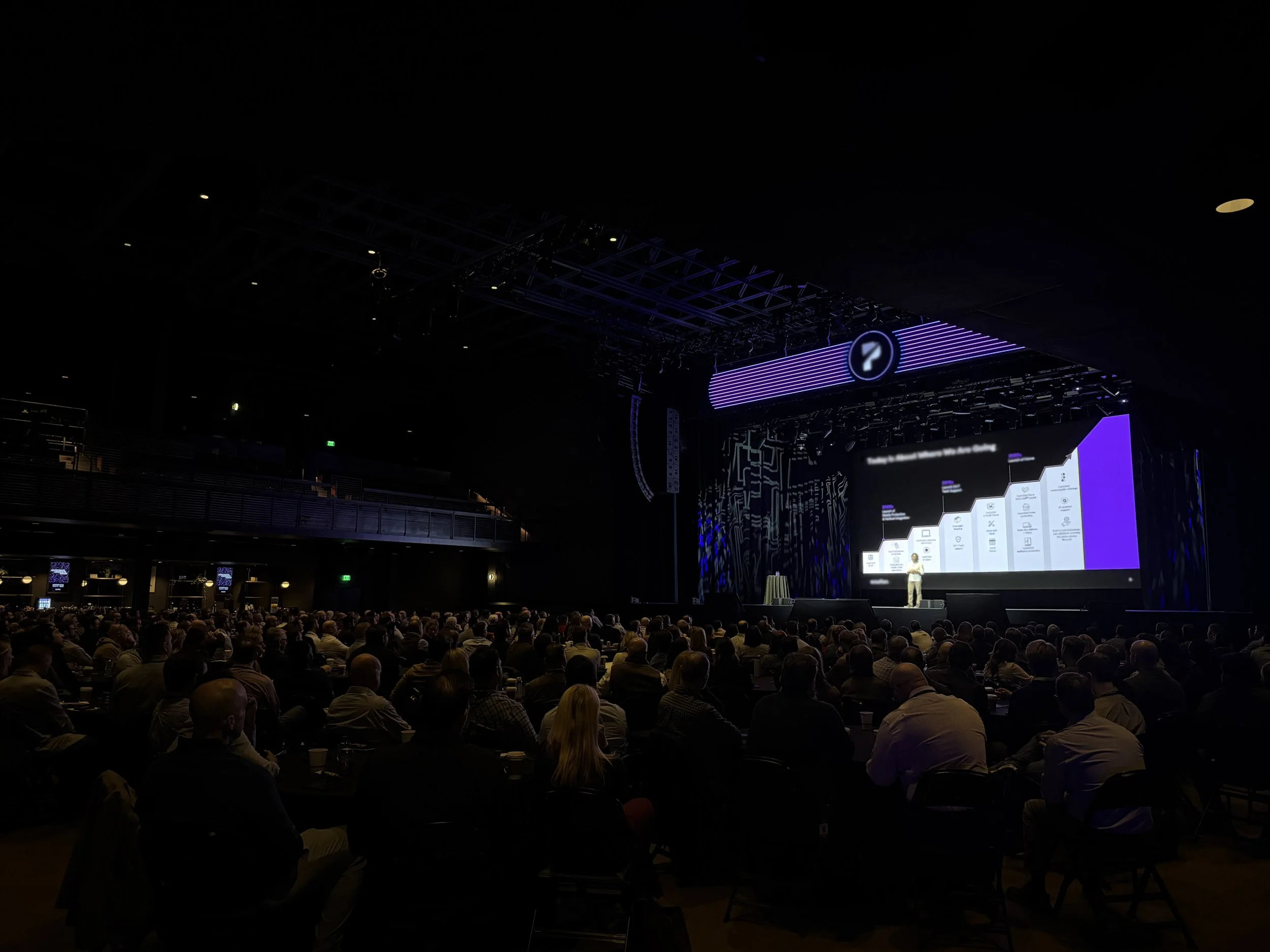 A large crowd seated in a conference hall listening to a presentation on stage. The stage has a large screen displaying graphics and text, with purple lighting accents.
