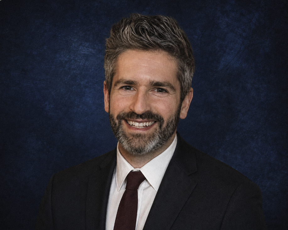 A smiling man with salt and pepper hair and a beard, dressed in a suit and tie, standing against a dark blue background.