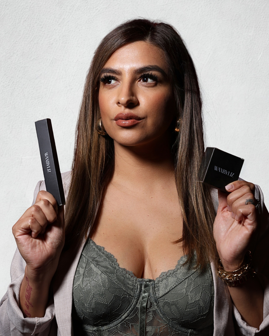 A woman with long brown hair holding black cosmetic boxes with 'WAXBYLIZ' branding, wearing a lace top and a blazer, standing against a plain white wall.