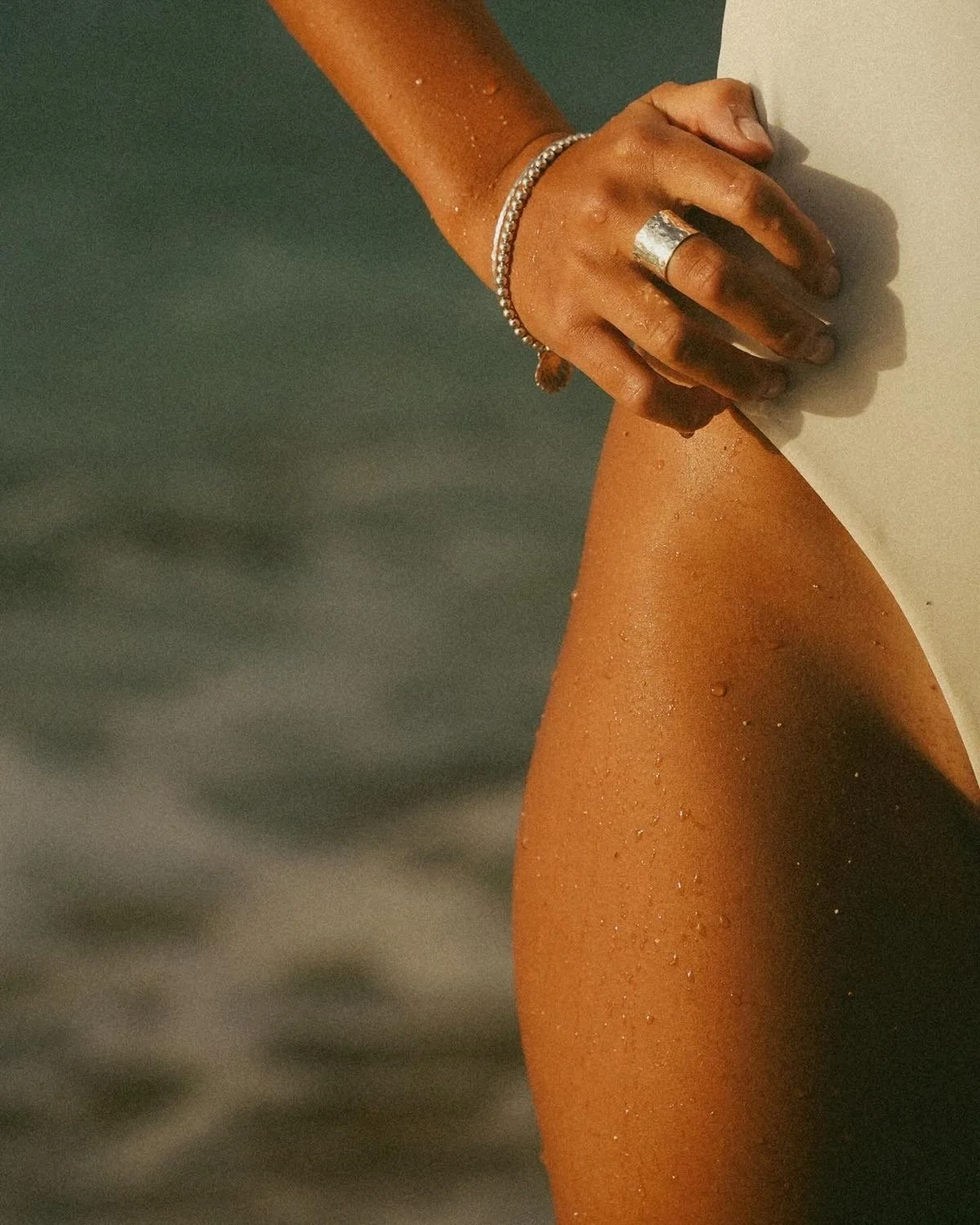 Close-up of a person's hand and leg by the water, with water droplets on the skin, wearing jewelry including a ring, bracelet, and a beaded bracelet.