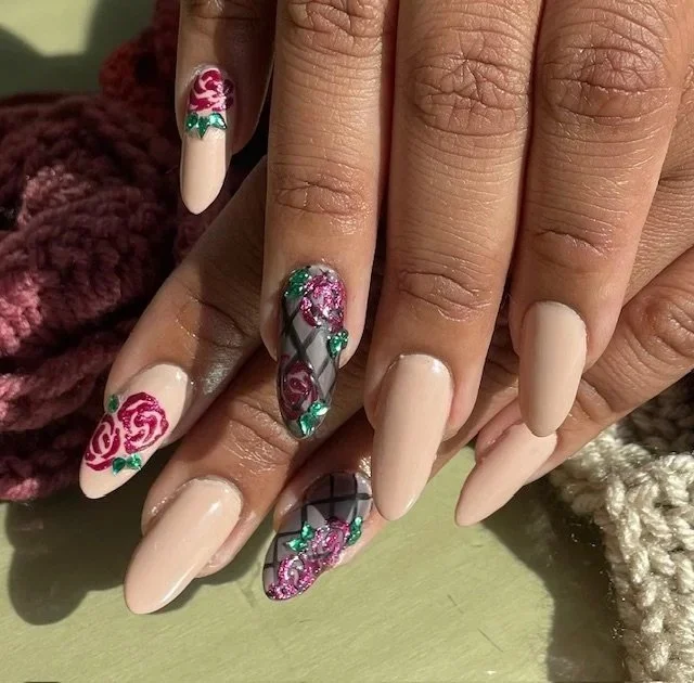 Close-up of hands with elaborately decorated acrylic nails featuring pink rose designs, green accents, and a diamond pattern on a neutral beige base.