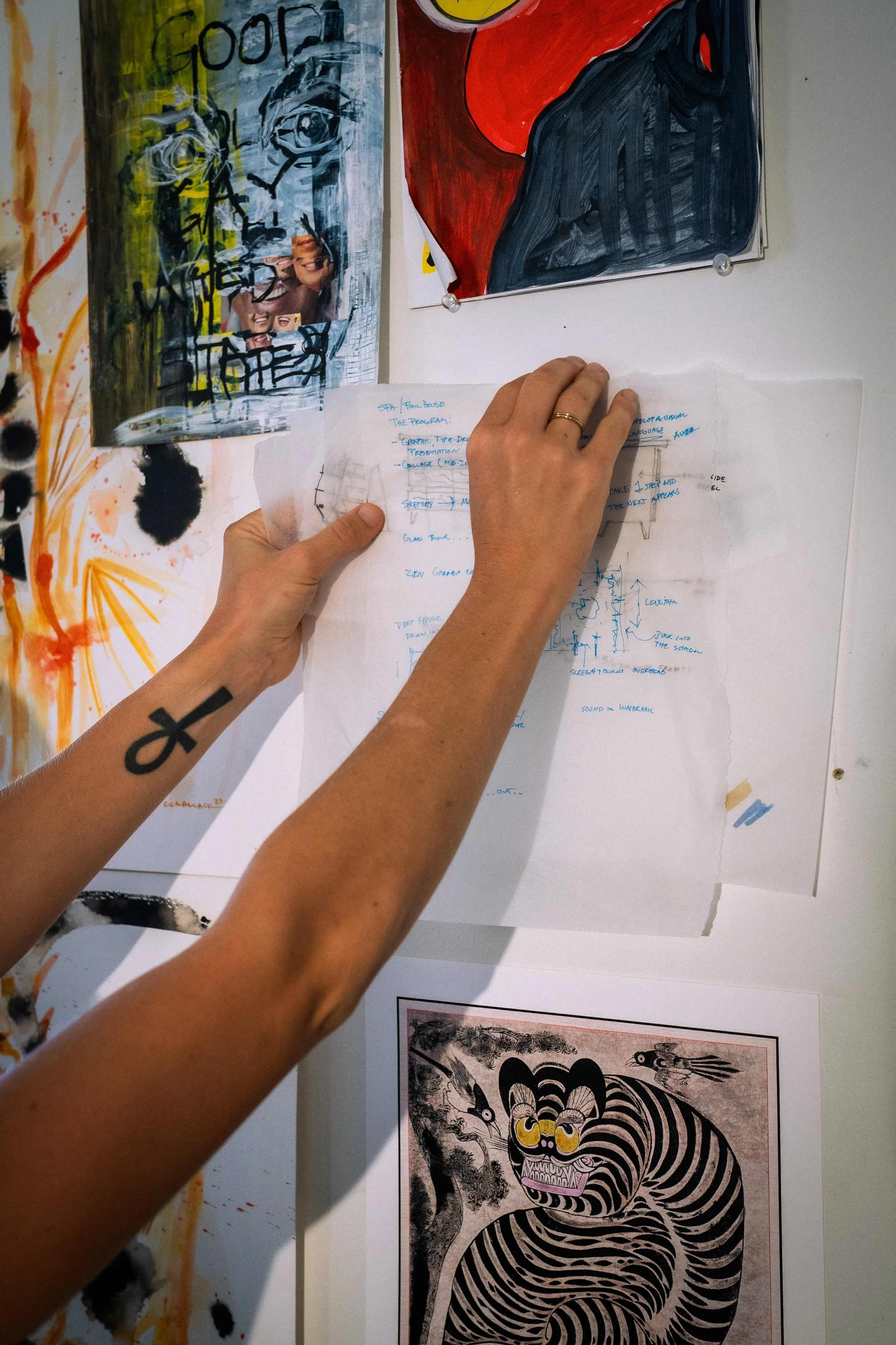 Artist and architectural designer, Caroline Wallace, taping a white sheet of paper with handwritten notes onto a wall, surrounded by colorful abstract artwork and posters, including an illustrated tiger at the bottom, and a different artwork above.