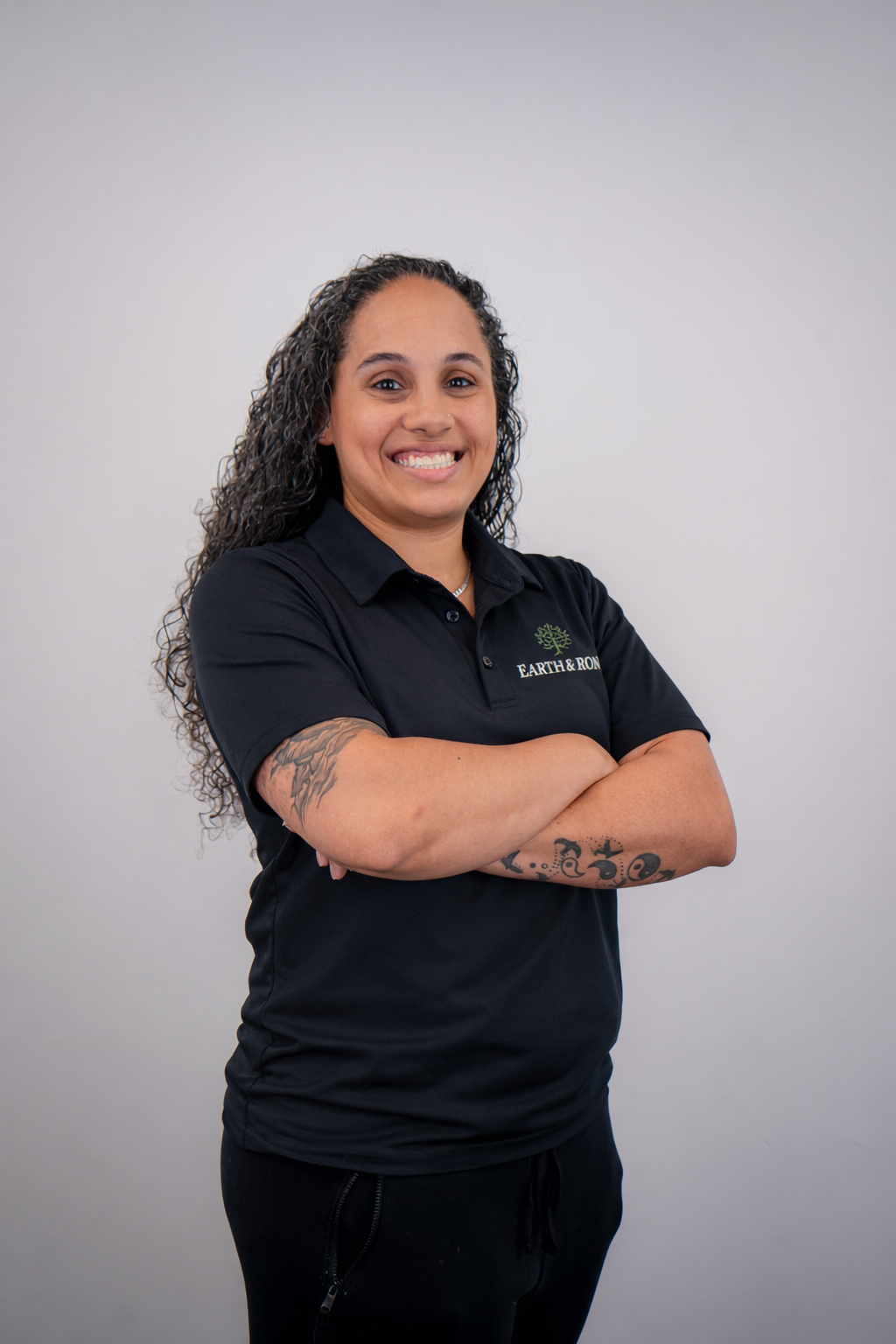 A woman with curly hair, tattoos on her arms, and a black polo shirt with the Earth & Rion logo stands with her arms crossed and smiles at the camera against a plain white background.