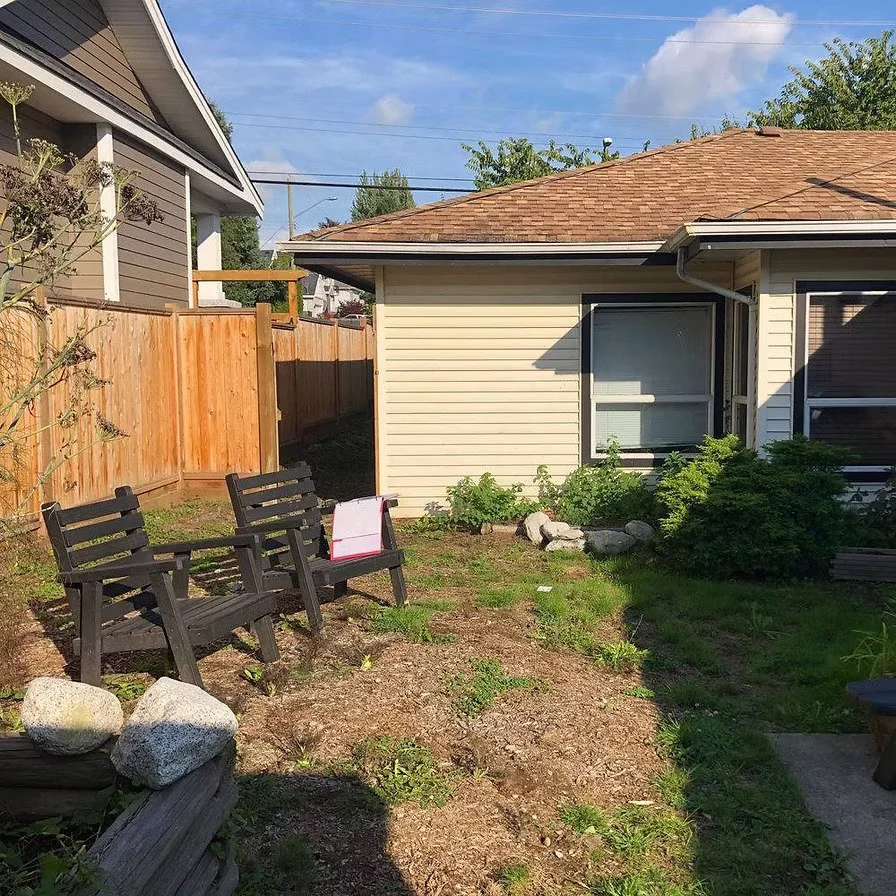 A backyard with two black wooden benches, some large rocks up against the benches, green plants and shrubs, a beige house with a window, a brown roof, a wooden fence, and a blue sky with some clouds.