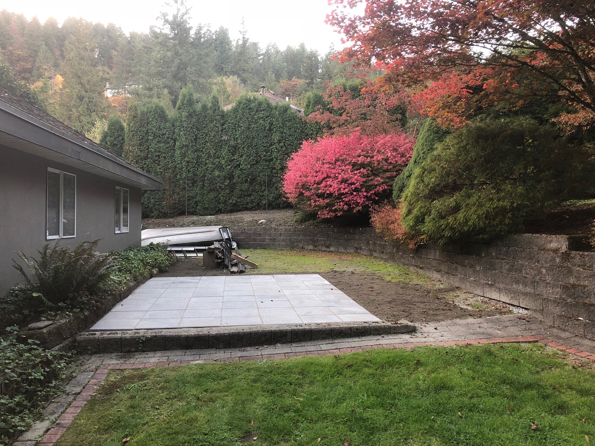 Backyard with a patio, green lawn, and colorful autumn trees including a pink-flowered shrub, with a house on the left and a stone retaining wall in the background.