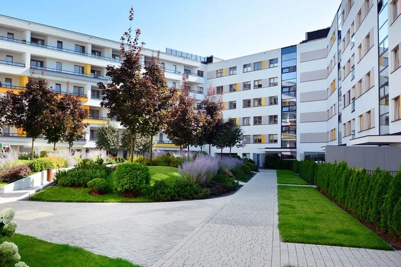 Modern apartment complex courtyard with paved walkways, lush green grass, trimmed bushes, and colorful trees, surrounded by white multi-story buildings.