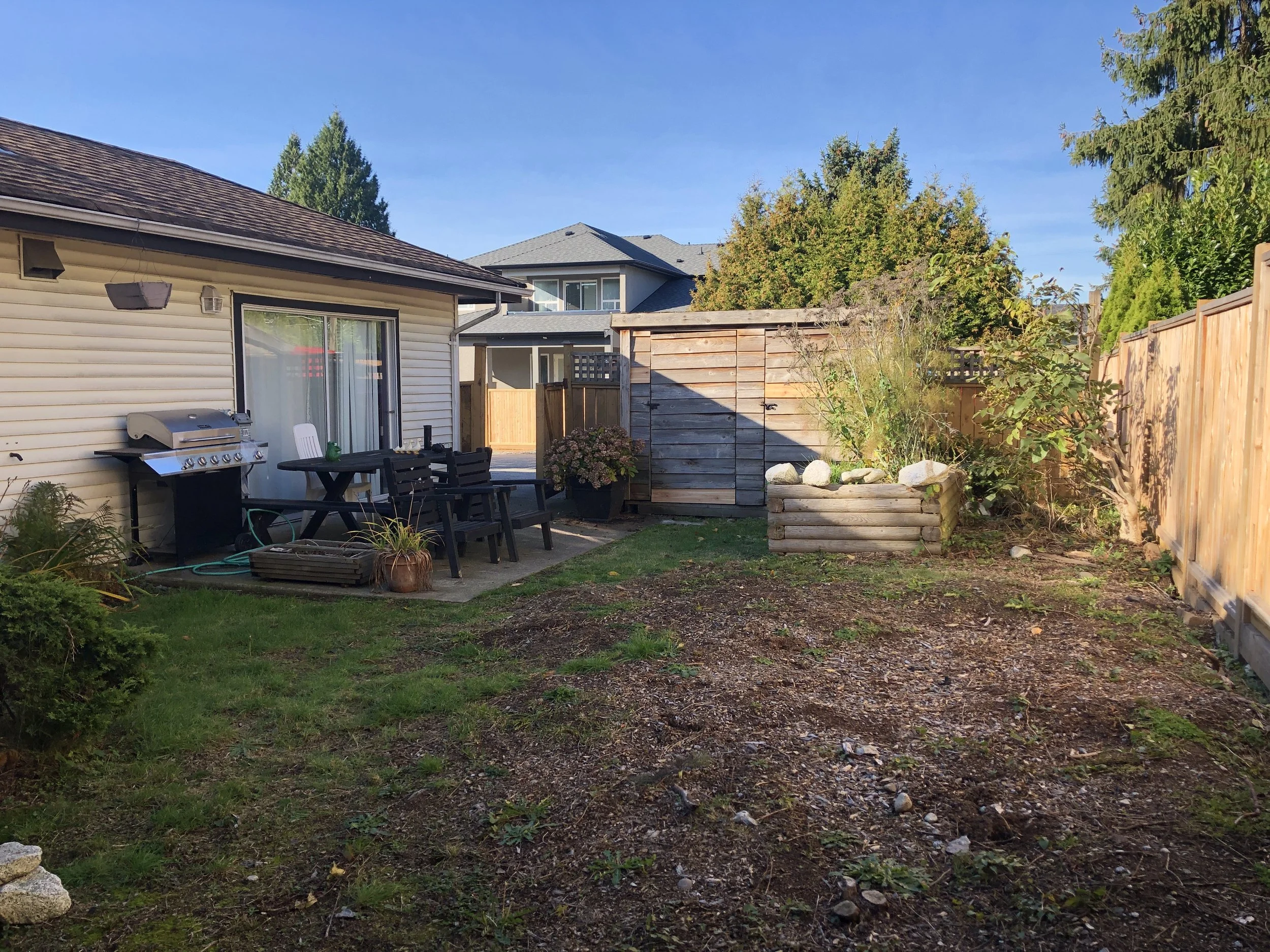 Backyard with patio table and chairs, grill, garden bed with rocks, bushes, trees, and wooden fence under clear sky.