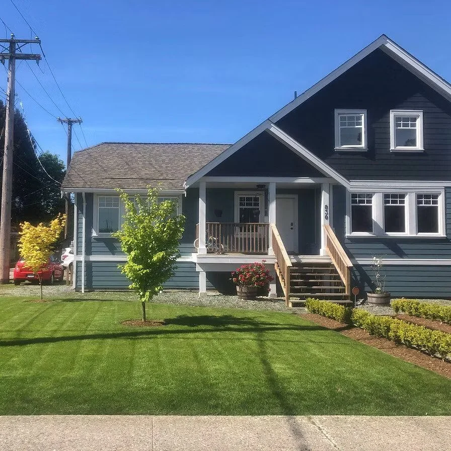 A blue house with a porch and stairs, a green lawn with small trees, and a clear blue sky.